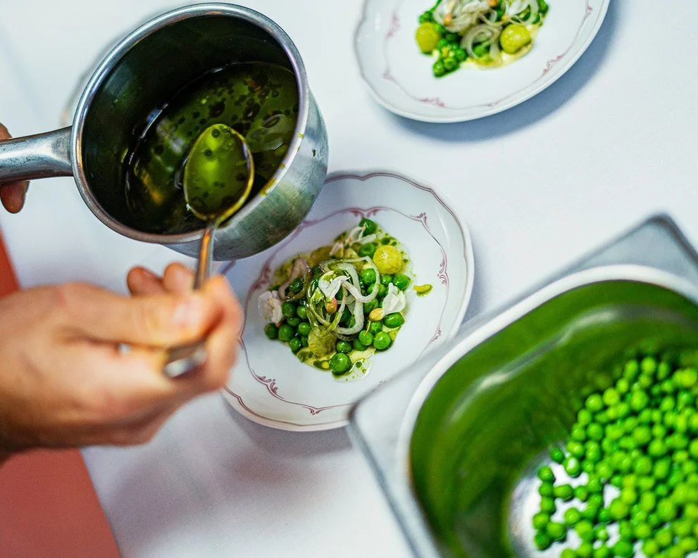 Person pouring green pea soup over a vegetable salad with peas, onions, and herbs in a white bowl with red trim, with additional bowls of peas and salad on a white table.