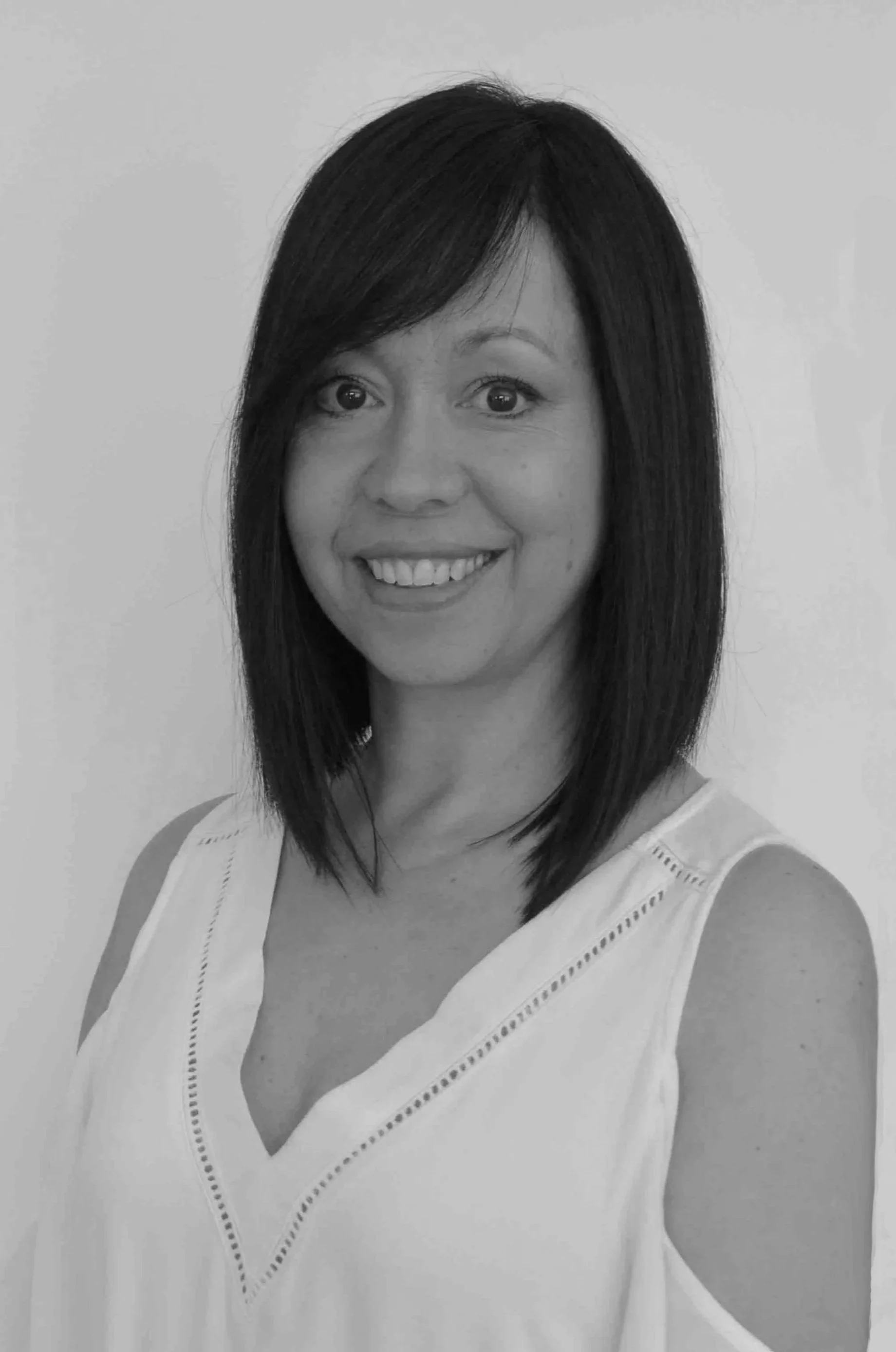 A woman with straight, shoulder-length dark hair, smiling, wearing a sleeveless white top with decorative stitching.