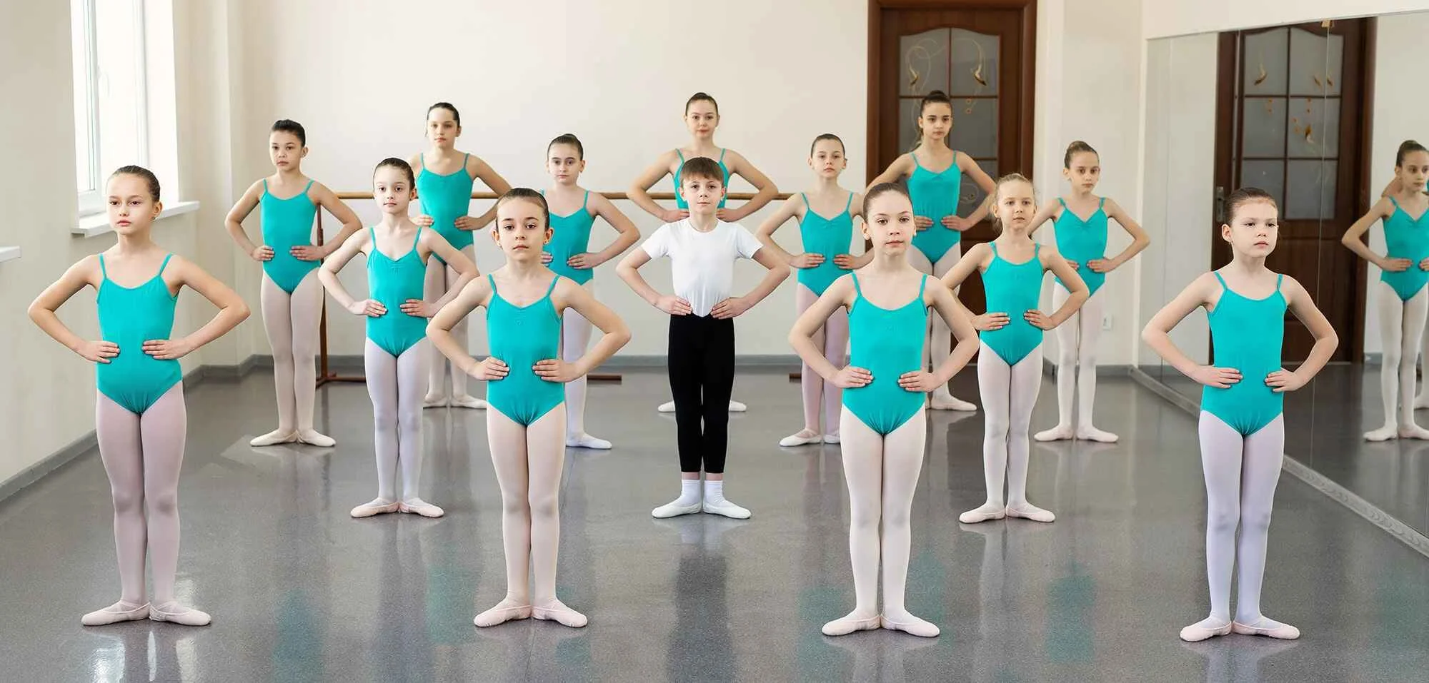 Group of young ballet dancers in turquoise leotards and white tights standing in a dance studio with mirrors, practicing ballet positions.
