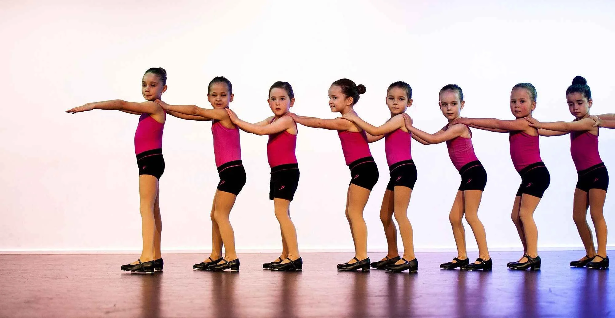 A line of young girls dressed in pink leotards and black ballet shoes practicing ballet on a stage.