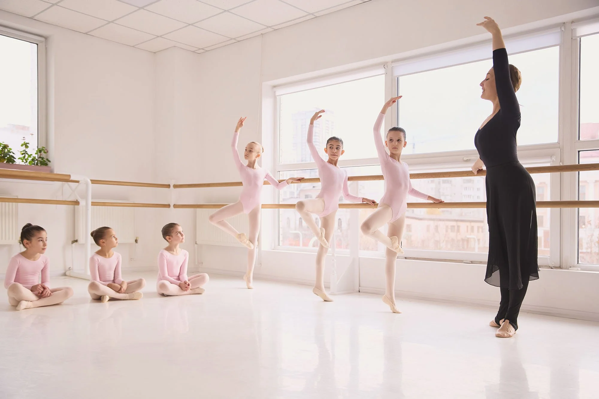 Ballet dance class with young girls and instructor in a studio with large windows, ballet barres, and natural light.
