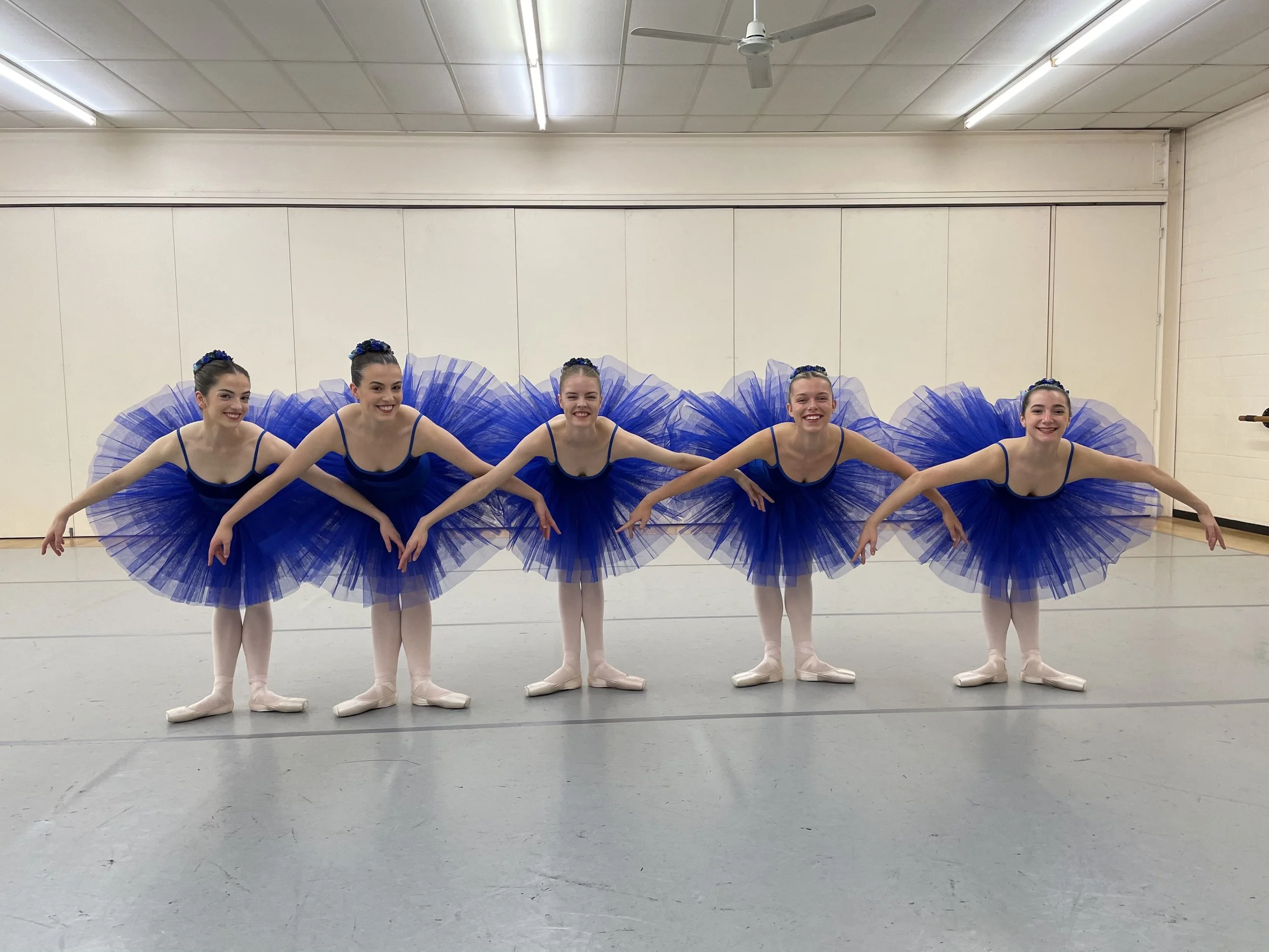 Five ballerinas in blue tutus and leotards are kneeling in a dance studio, holding hands and smiling in a line.
