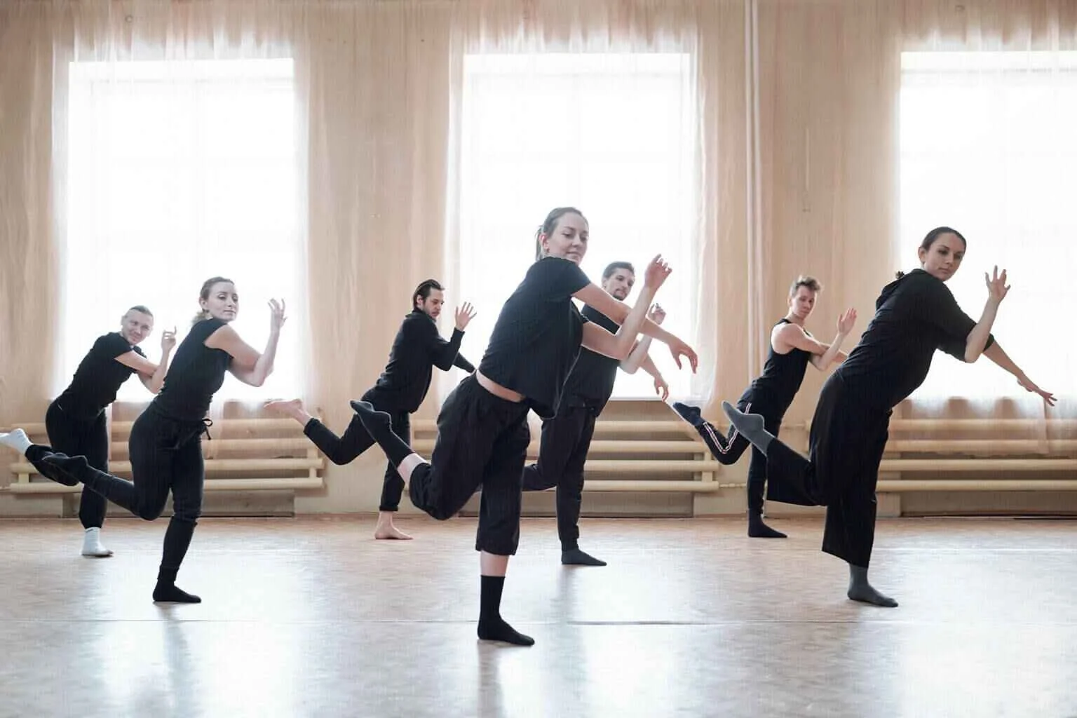 Group of seven dancers practicing in a spacious dance studio with large windows and light curtains.