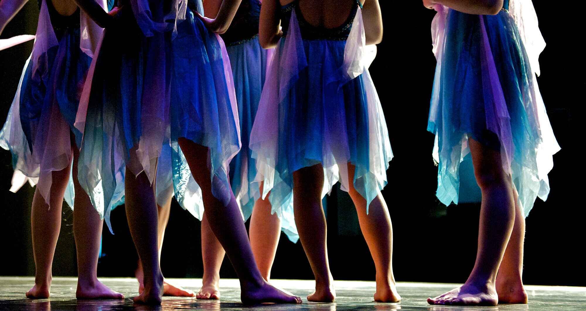 Ballet dancers in colorful tutus standing in a circle on stage, viewed from below with stage lights creating a silhouette effect.