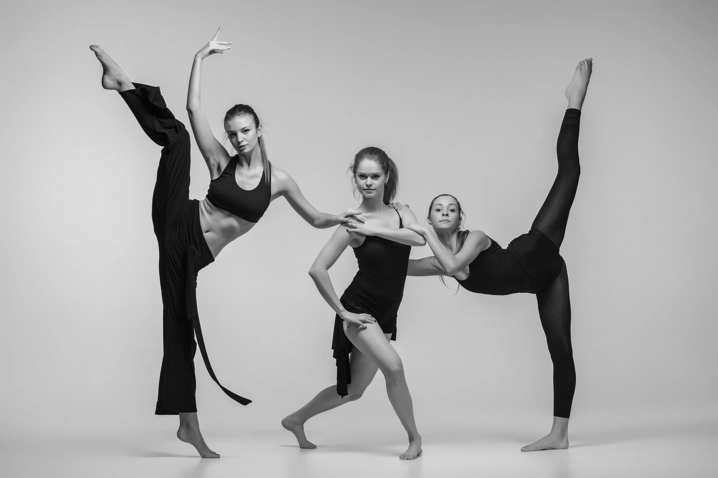 Black and white photo of three young women in dance poses, standing on one leg with the other leg extended, and arms in elegant positions against a plain background.
