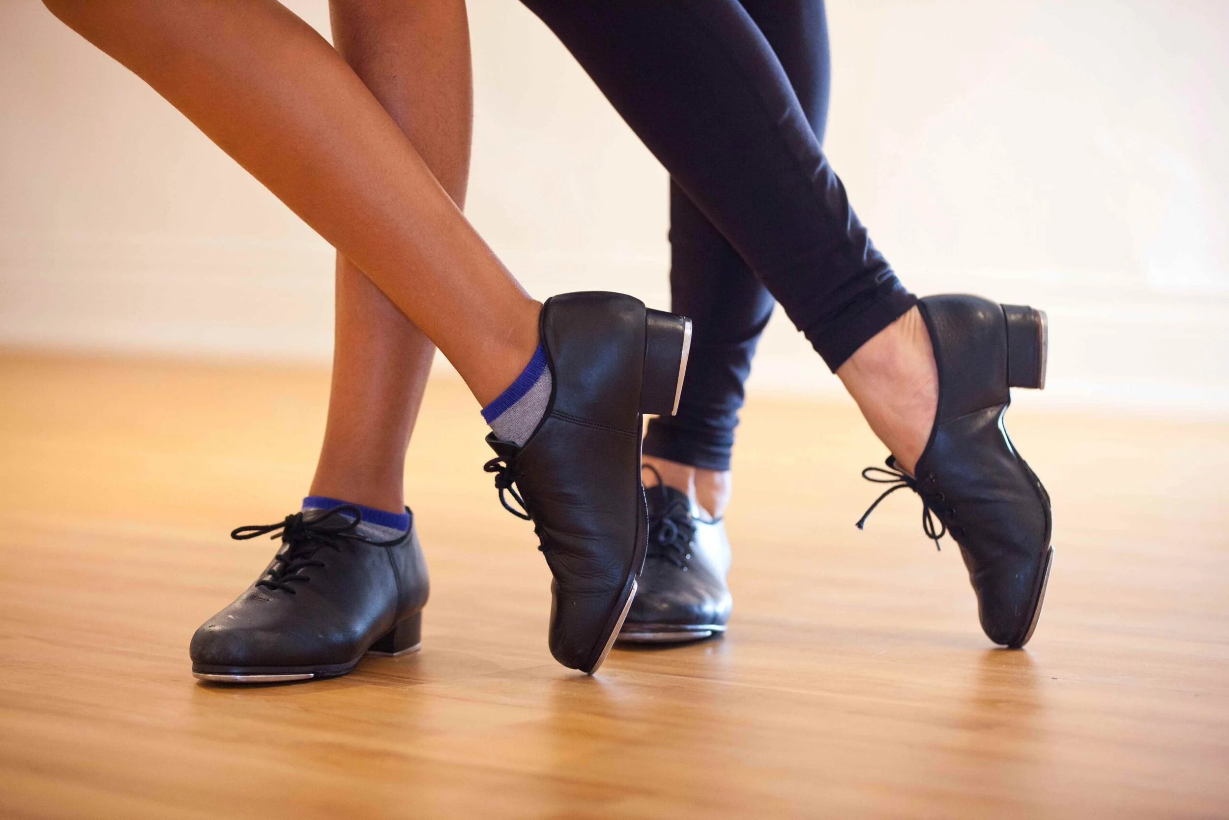Two people wearing black dance shoes with heels, black pants, and socks, dancing closely on a wooden floor.