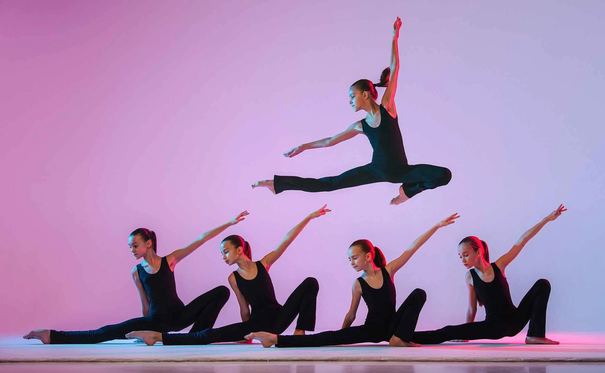 A group of young female dancers performing a choreographed dance routine on stage, with one dancer leaping mid-air above the others who are sitting or in a pose.