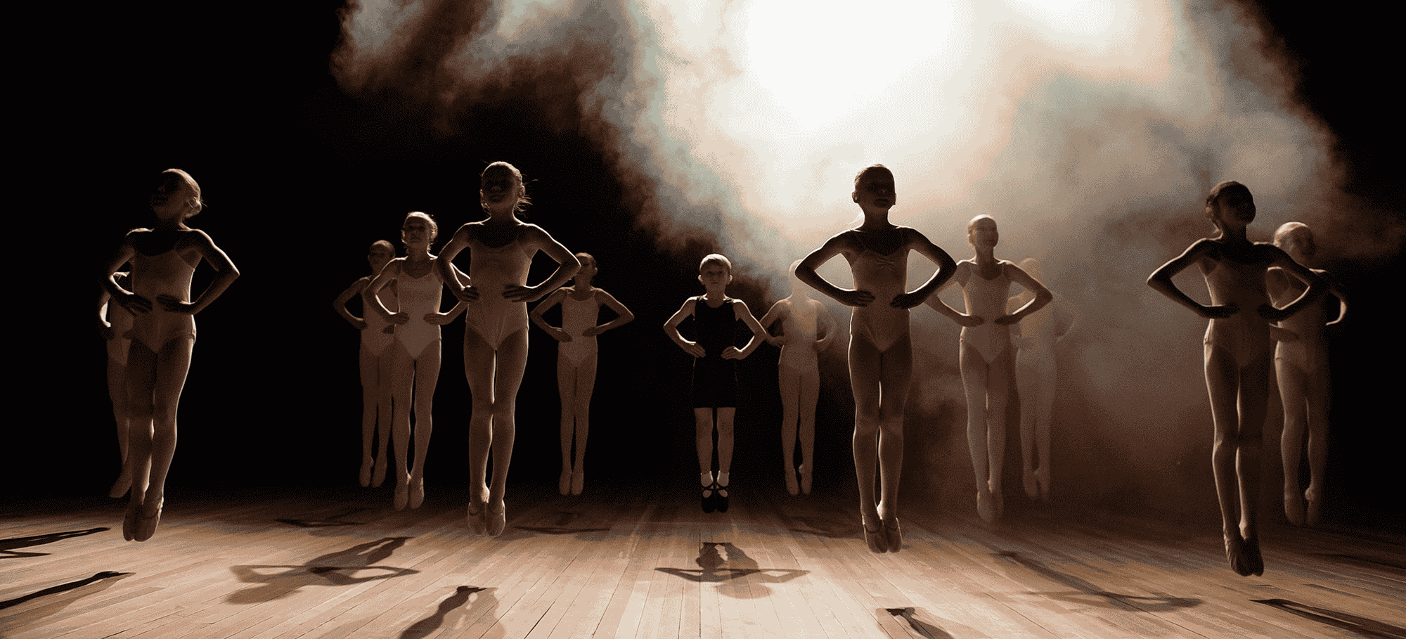 Group of ballet dancers, mostly young girls, practicing on stage with a foggy background and dramatic lighting.