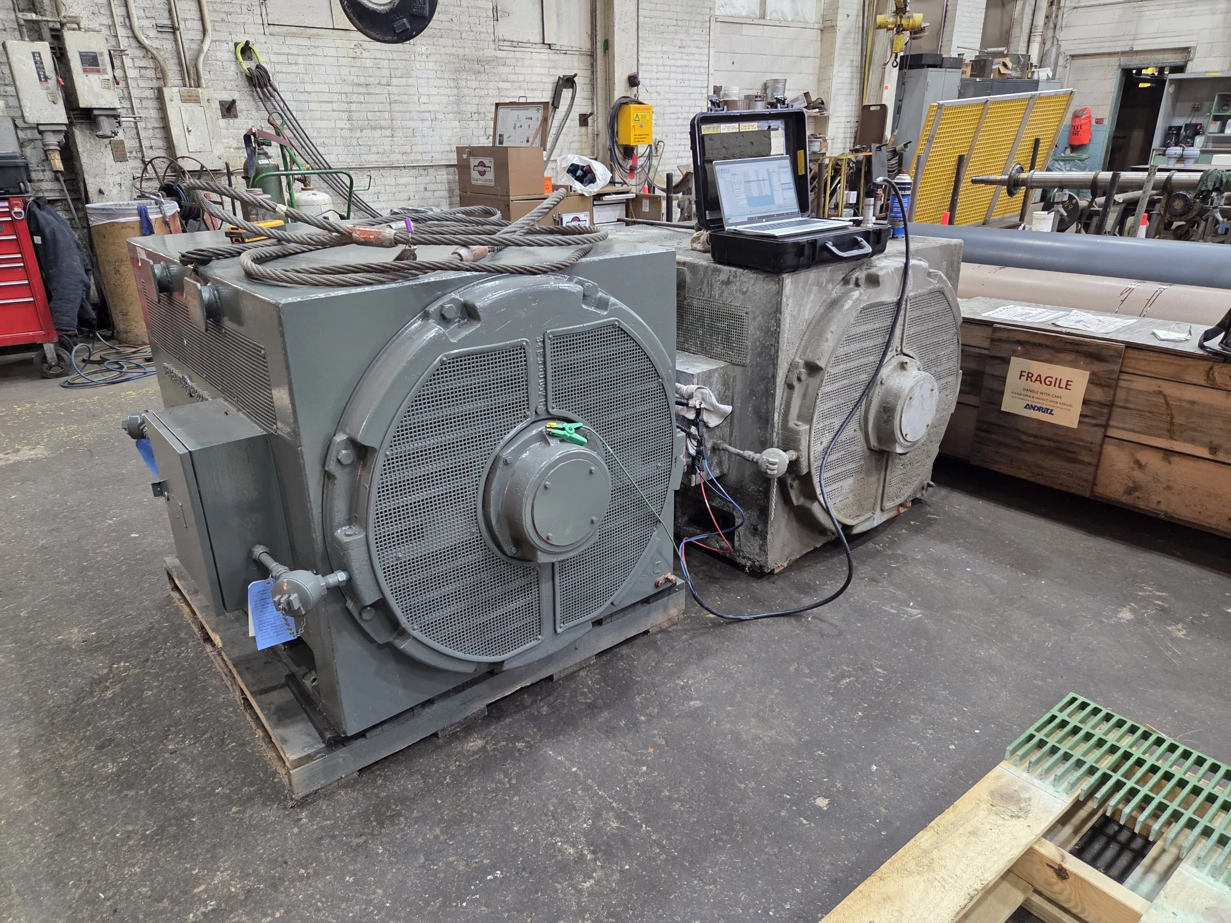 Two large industrial transformers with a laptop and testing equipment on top, inside a factory warehouse with tools and equipment in the background. 2300 volt ac motors. Offline testing. Comparing two motors. Baseline testing.