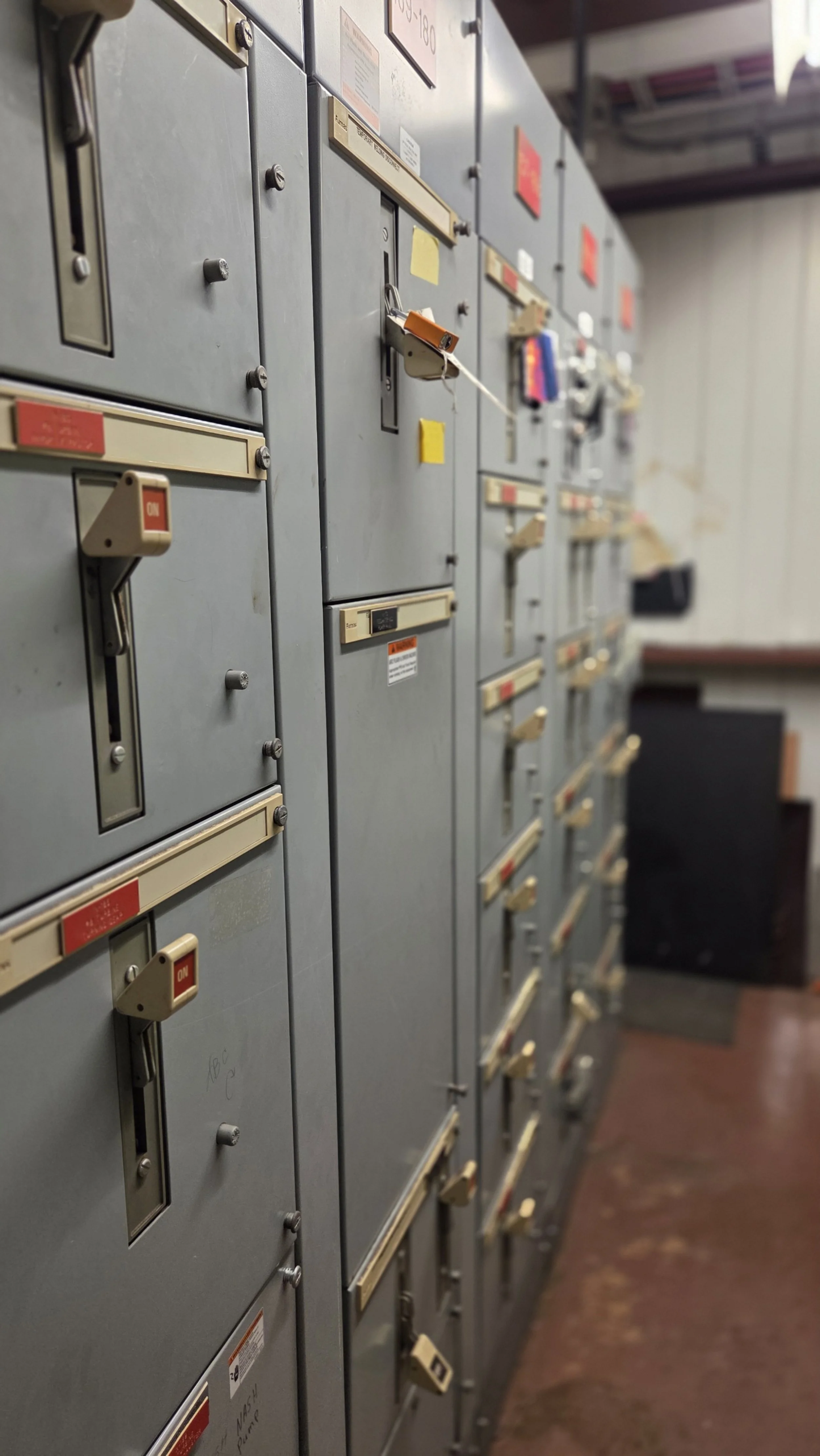 Row of gray electrical or utility lockers with various switches, locks, and labels, some with keys or padlocks attached, in an industrial or utility room.