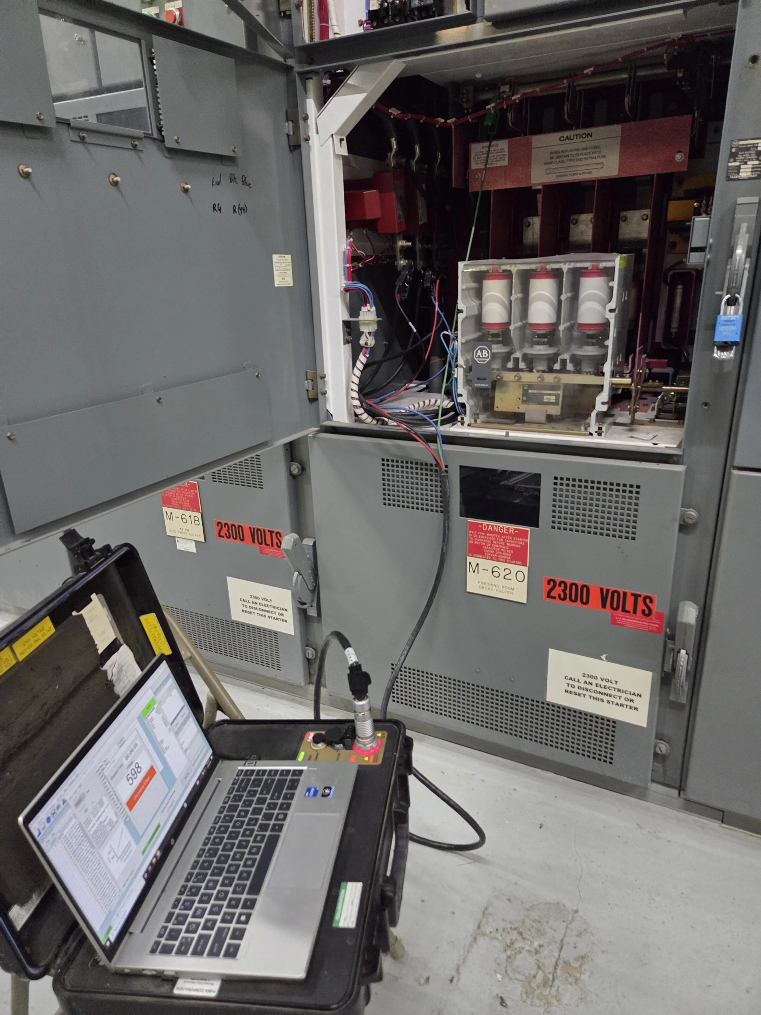 Open electrical cabinet with exposed components and wiring, connected to a laptop computer on a portable workbench, used for testing or diagnostics, with warning labels indicating high voltage.