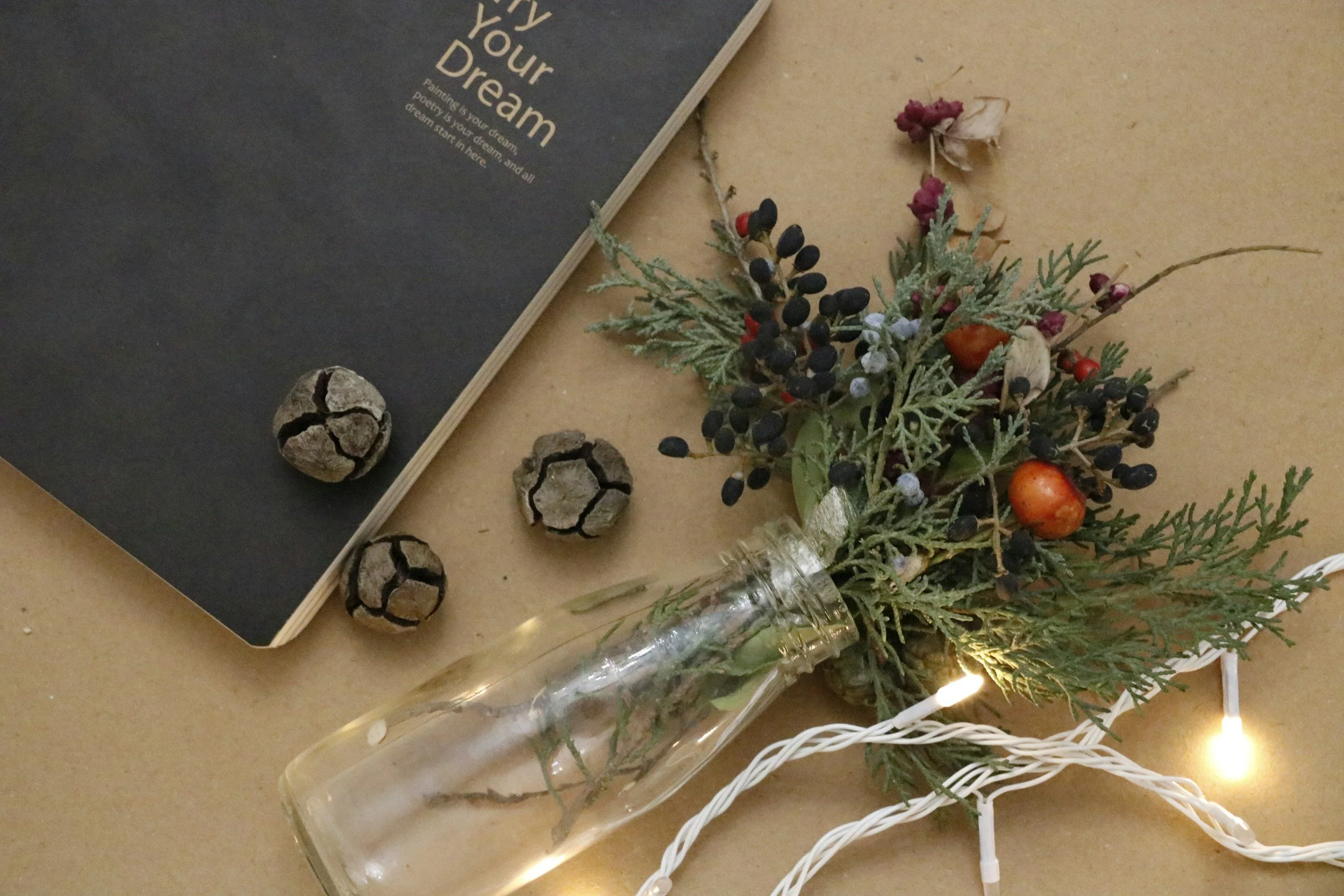 A flat lay of a black notebook with celestial text, three small decorative seed pods, a glass bottle with greenery, berries, and twinkle string lights on a tan surface.