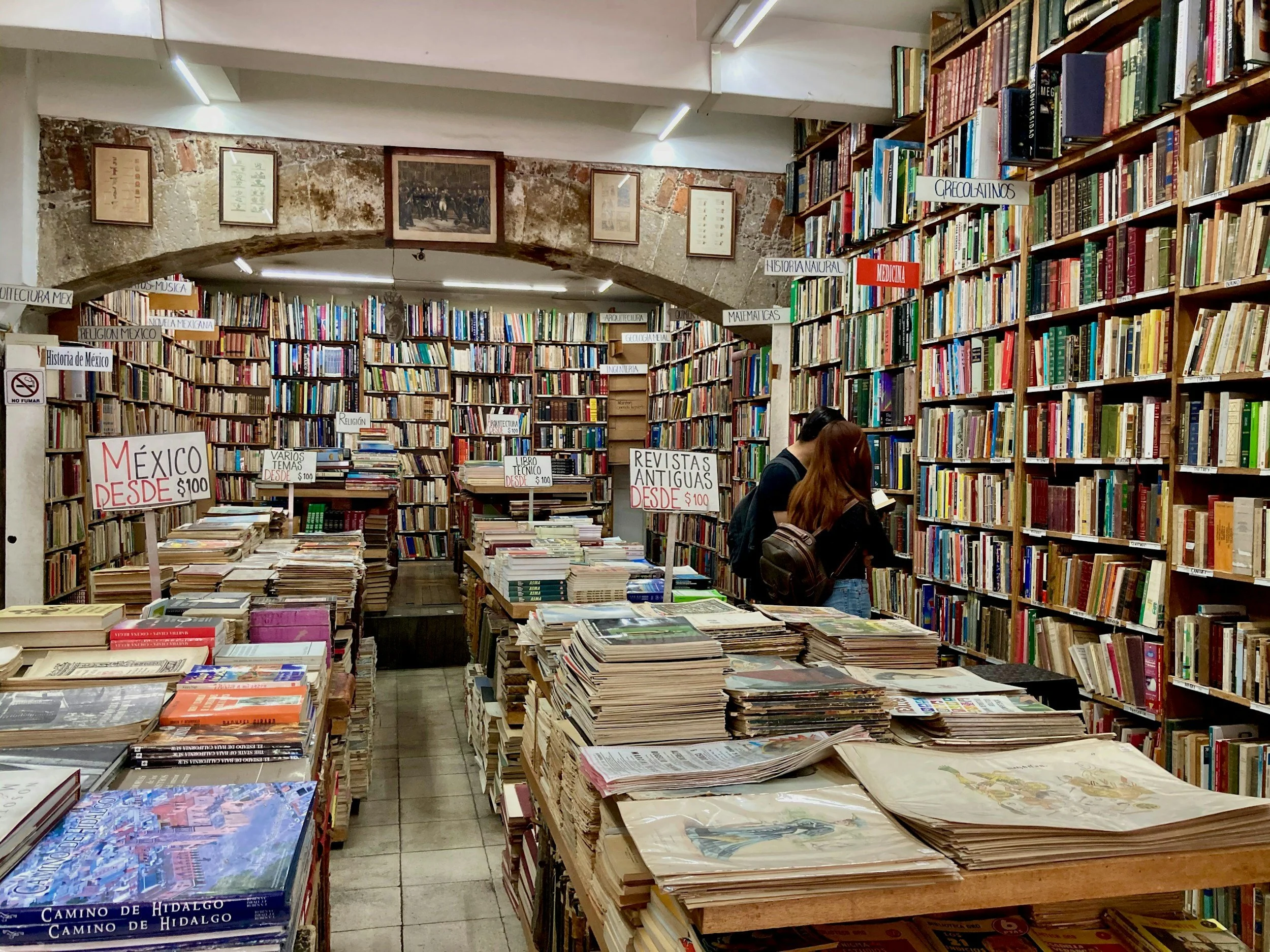 Interior of a bookstore with shelves filled with books, signs indicating sections like 'México', 'Revistas Antiguas', and others, and two people browsing the shelves.