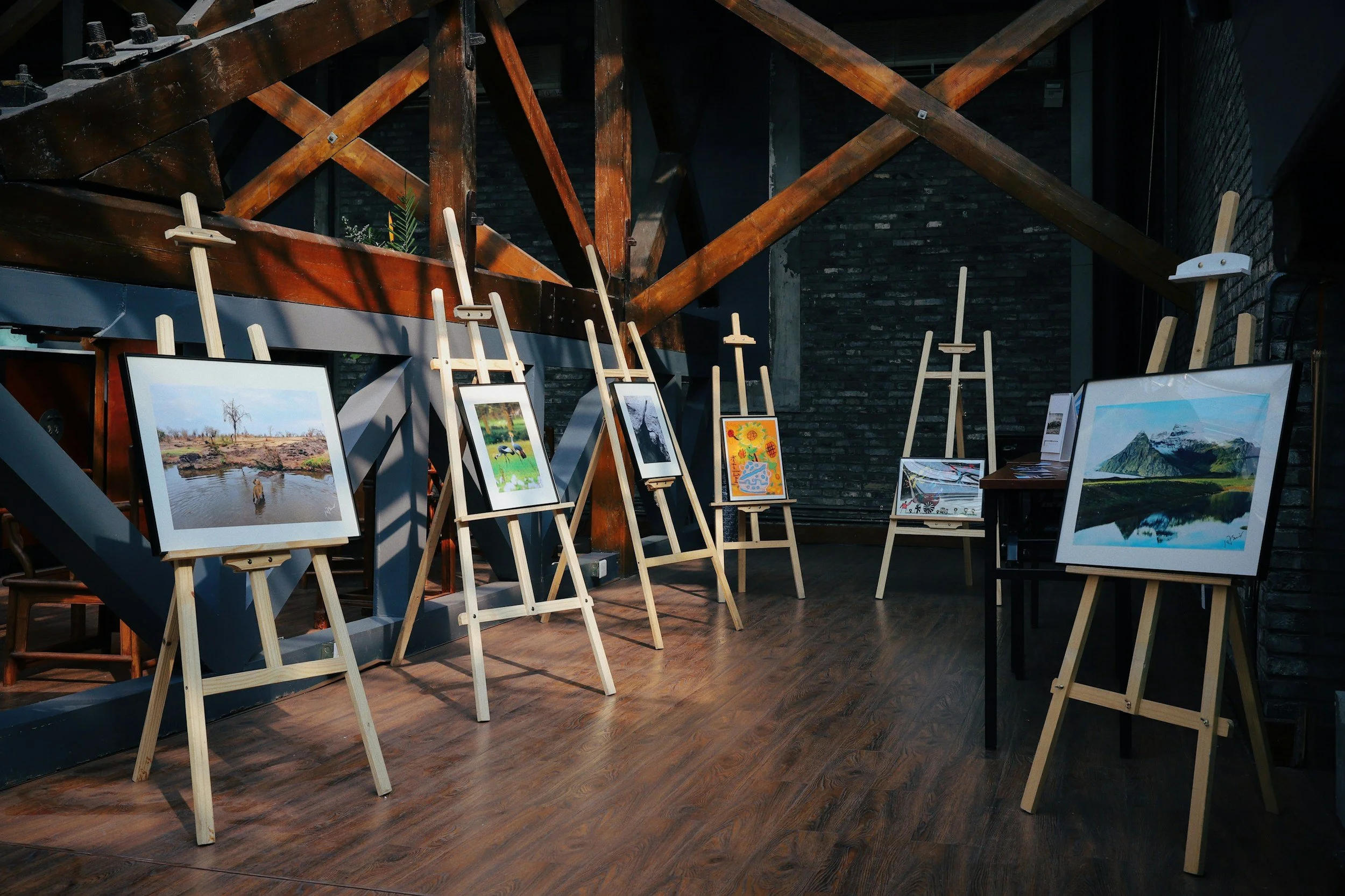 Art gallery with framed landscape and nature paintings on wooden easels, inside a room with dark brick walls and wooden beams.