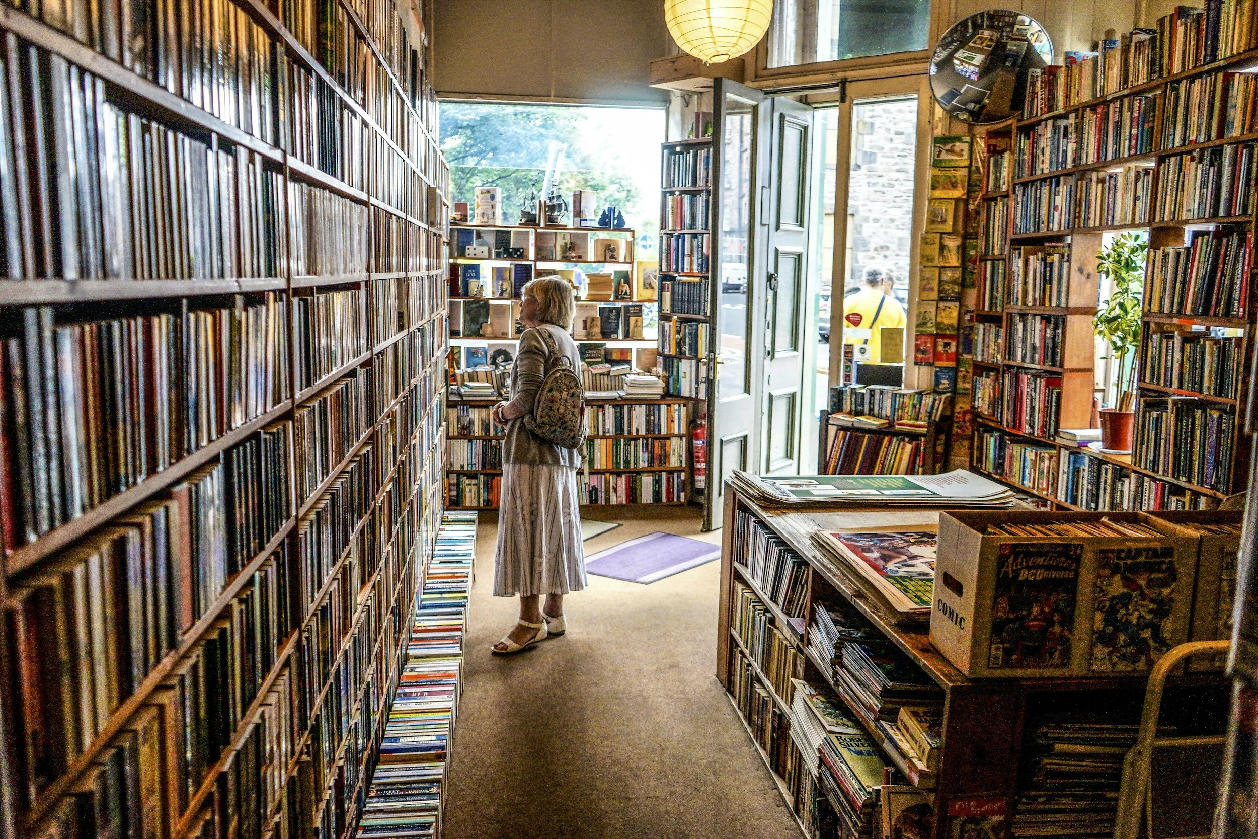 A woman in a skirt and sandals with a backpack browsing books inside a cozy used bookstore.