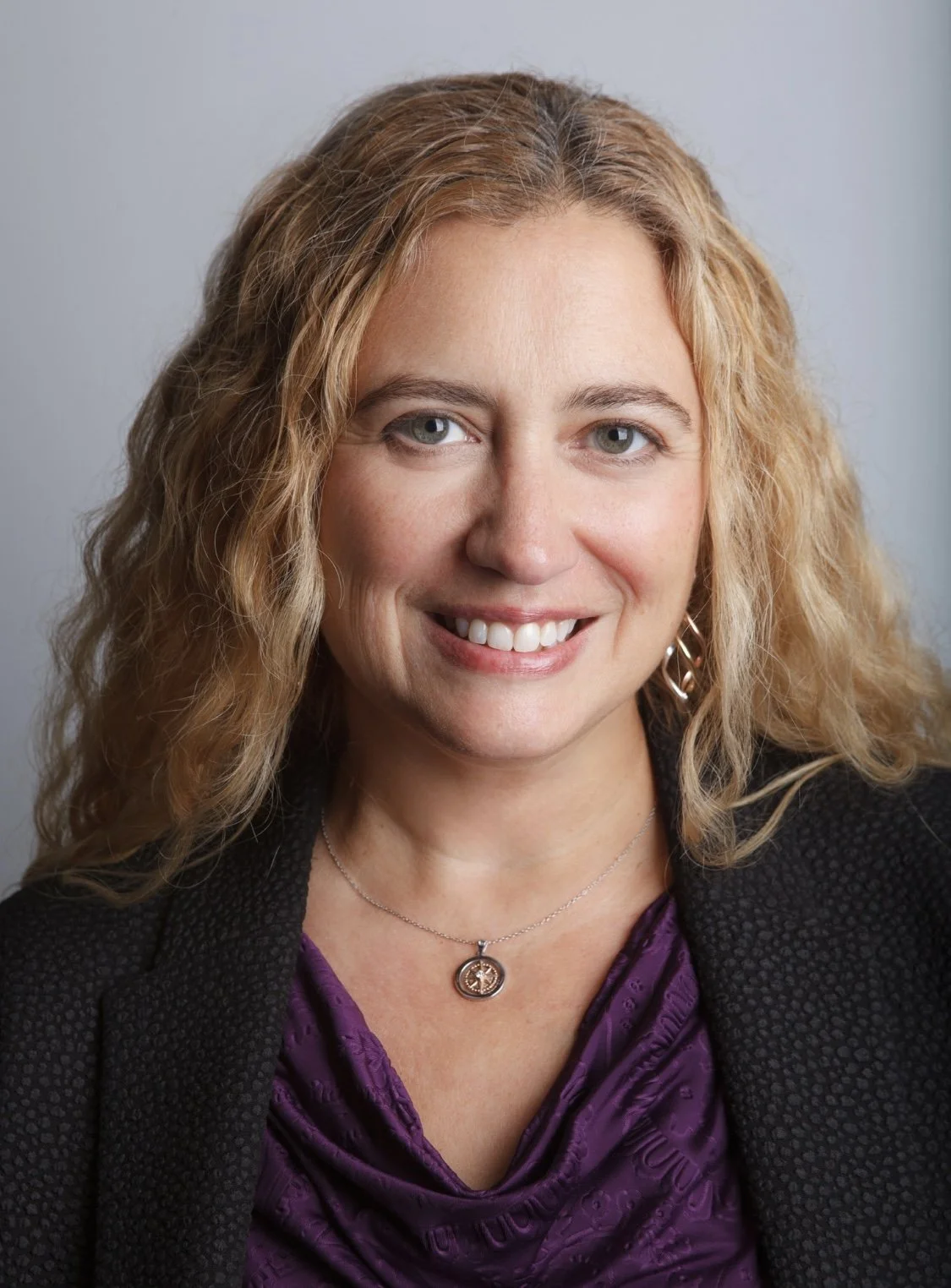Close-up portrait of a woman with blonde, curly hair, wearing a black blazer over a purple top, a silver necklace, and earrings, smiling at the camera against a plain gray background.