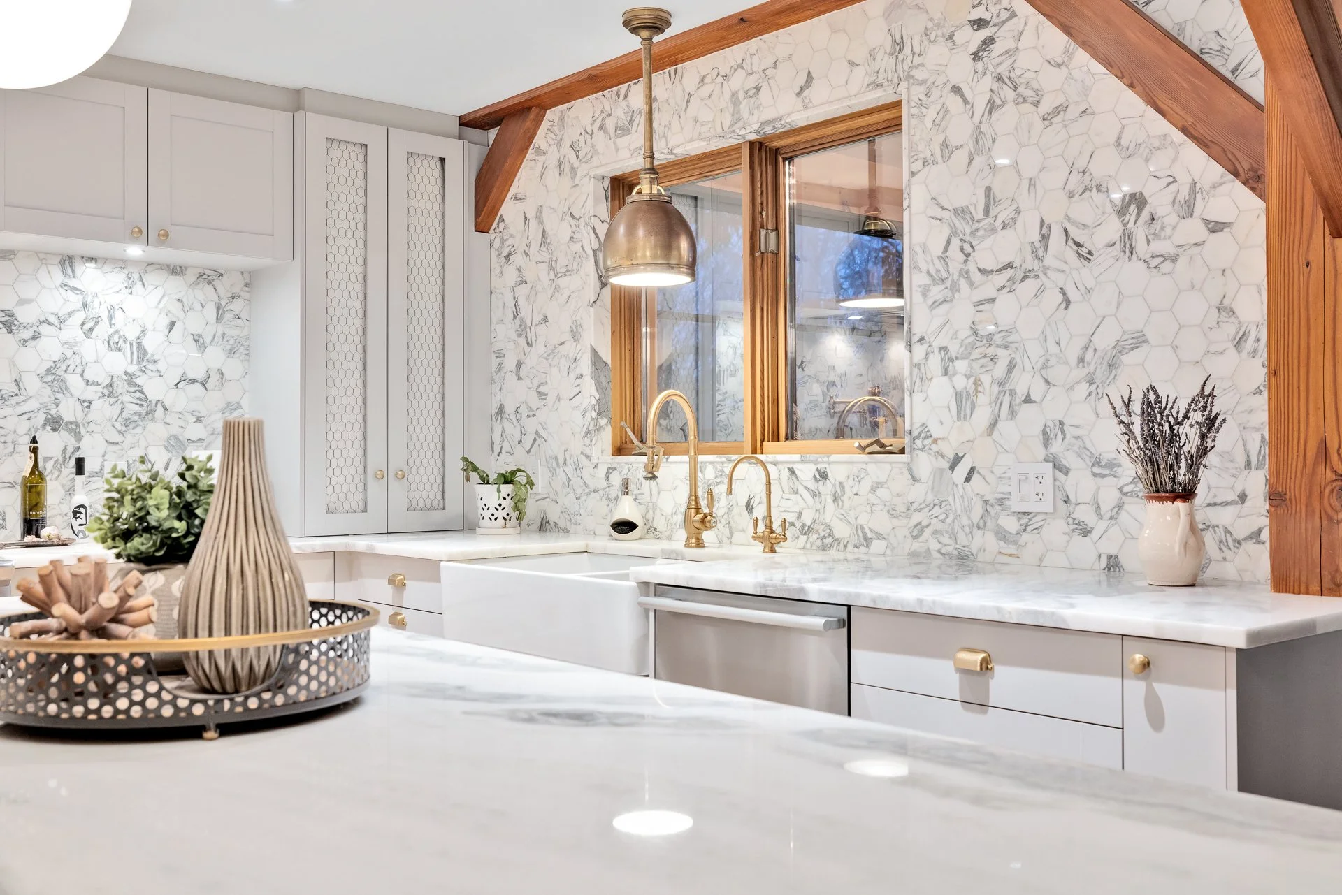 A modern kitchen with white cabinets, marble countertops, and a marble backsplash. There are gold fixtures, a decorative vase with dried lavender, and a window with a wooden frame.