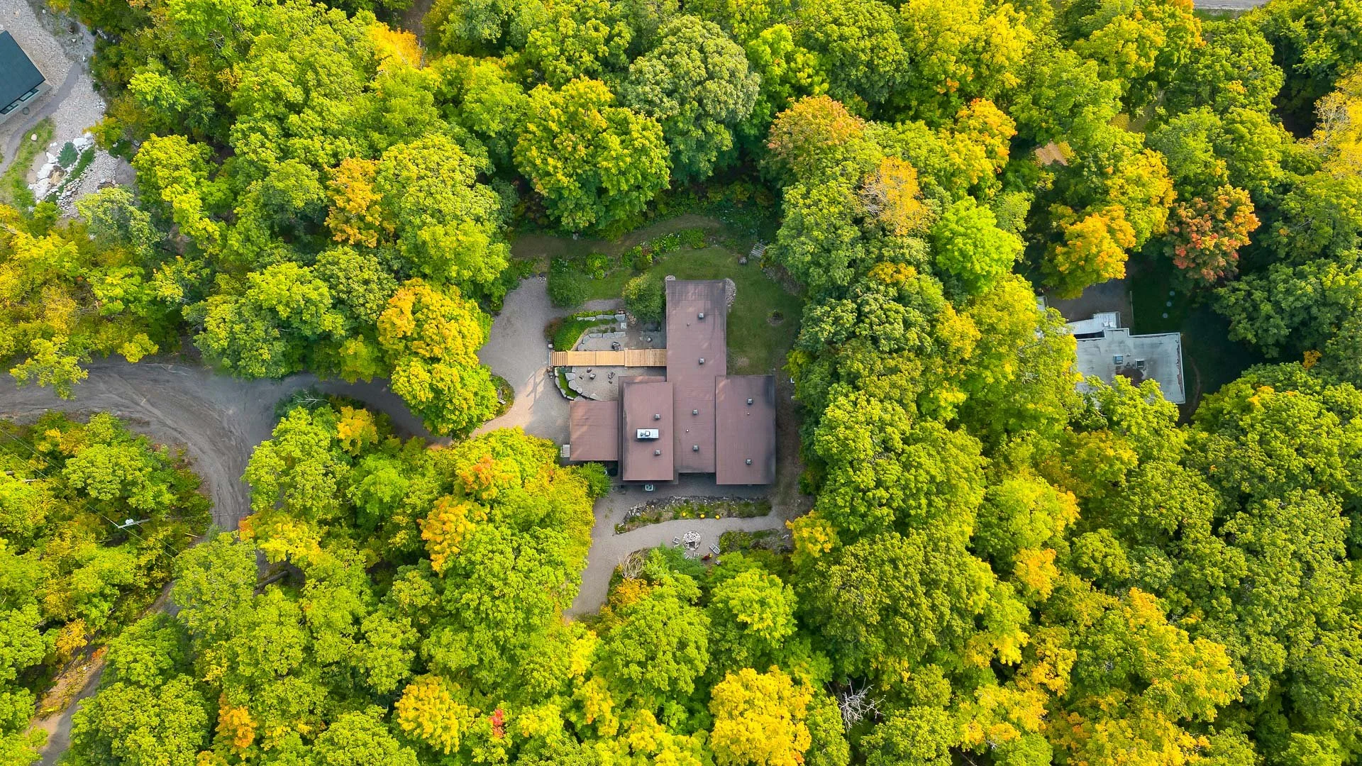 Aerial view of a house surrounded by dense green and yellow trees, with a gravel driveway and outdoor seating area.