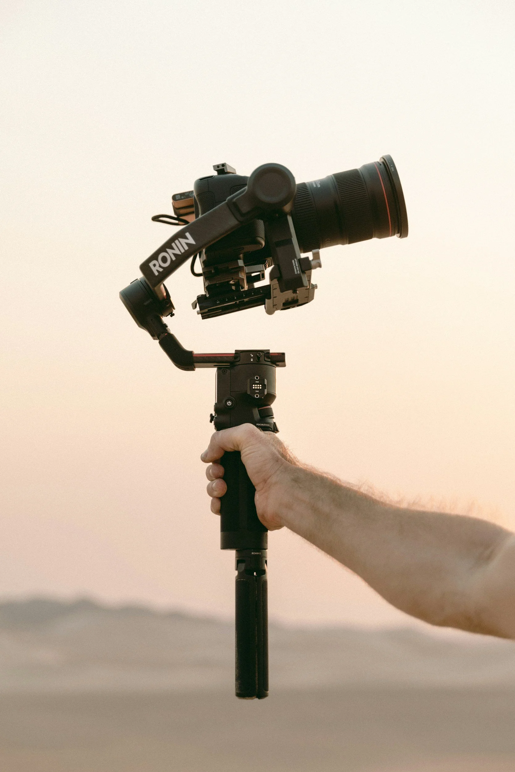 A person holding a camera stabilizer with a camera attached during sunset with a clear sky and distant mountains in the background.
