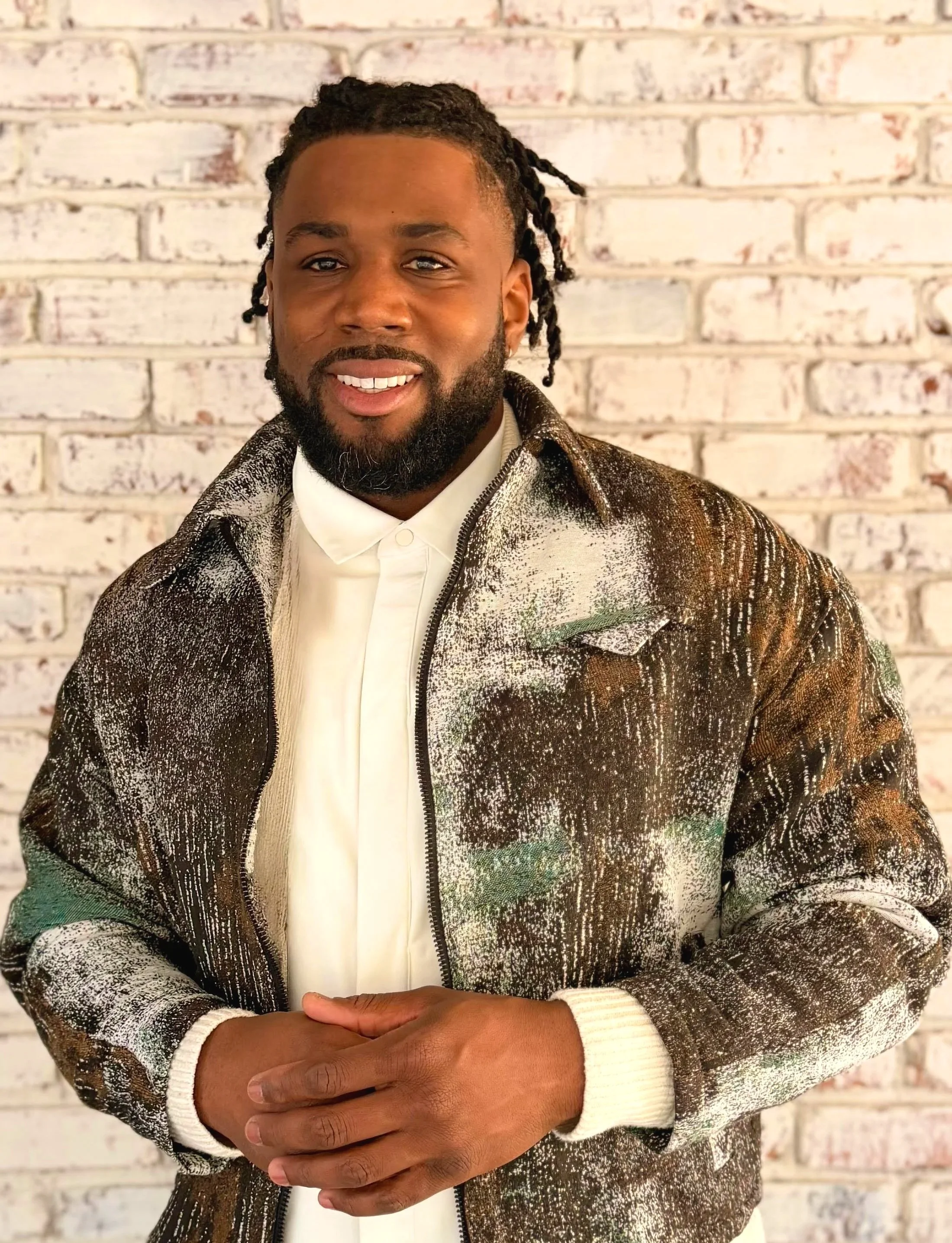 A man with dreadlocks and a beard wearing a brown, green, and white patterned zip-up jacket over a white shirt, standing in front of a white brick wall.
