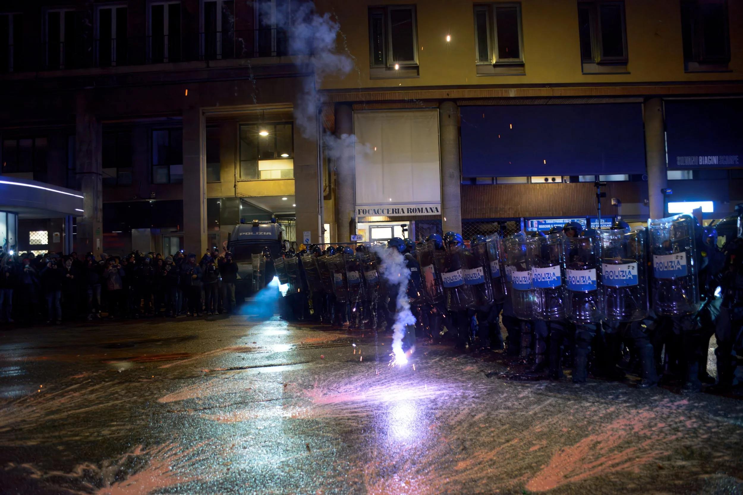 Bologna, Italy – November 21, 2025: Riot police hold the line with shields raised as tear gas drifts through the rain-soaked street, moments before a firework explodes on the wet pavement in front of their formation during clashes over the Virtus–Mac