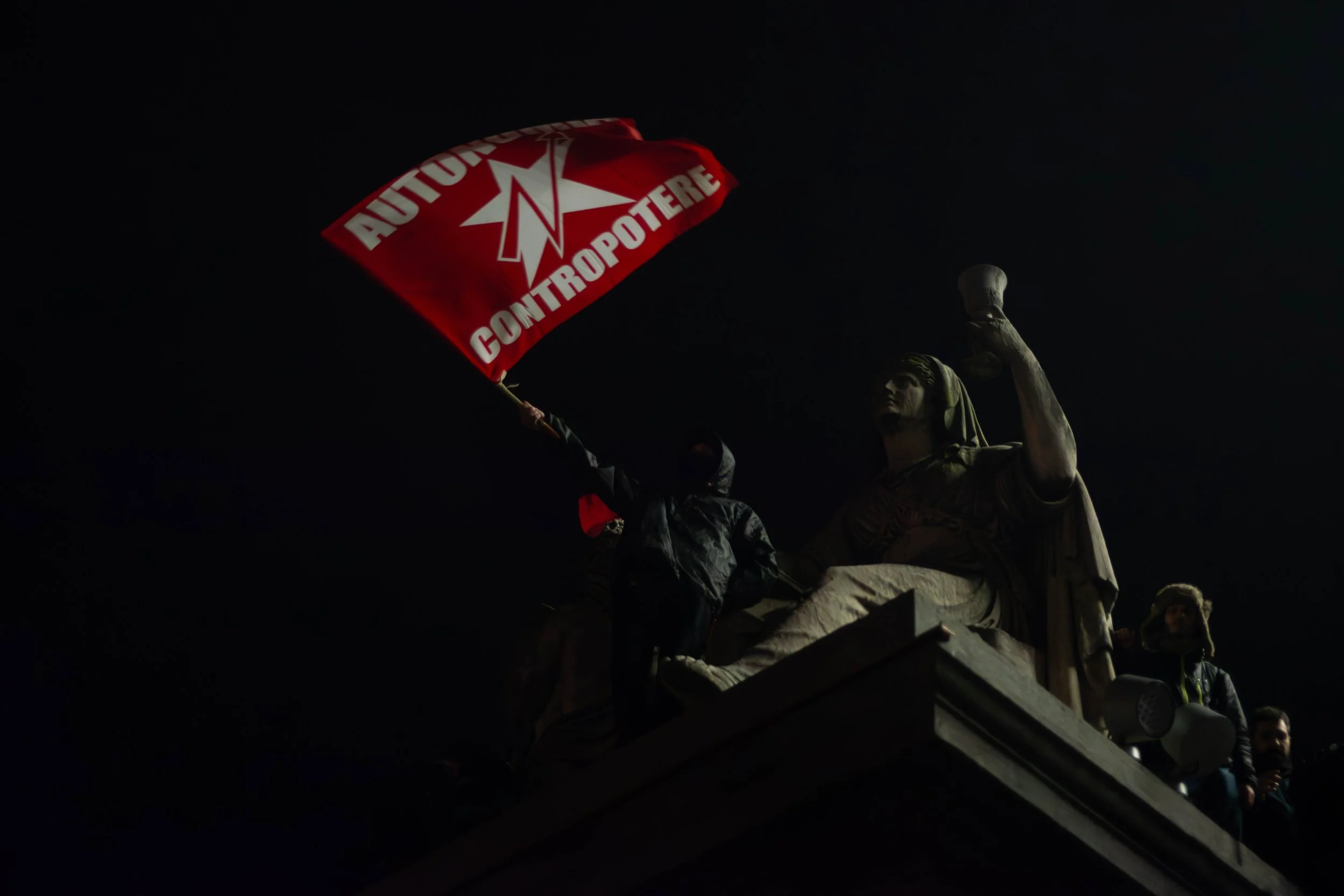 Turin, Italy – December 20, 2025: A hooded protester waves a red flag from the top of a monument at night, turning the statue into a symbol of resistance after the eviction of the Askatasuna social center.