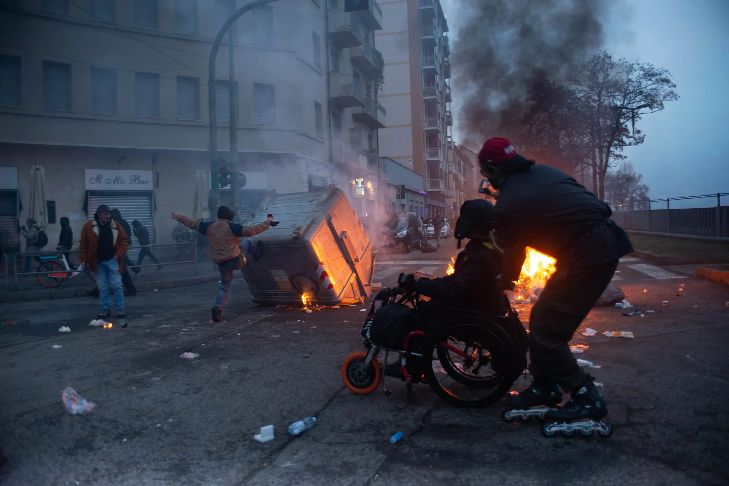 Turin, Italy – December 20, 2025: A protester kicks an overturned trash container as others move past burning debris, including a demonstrator in a wheelchair being pushed through the smoky street during clashes over the Askatasuna social center.