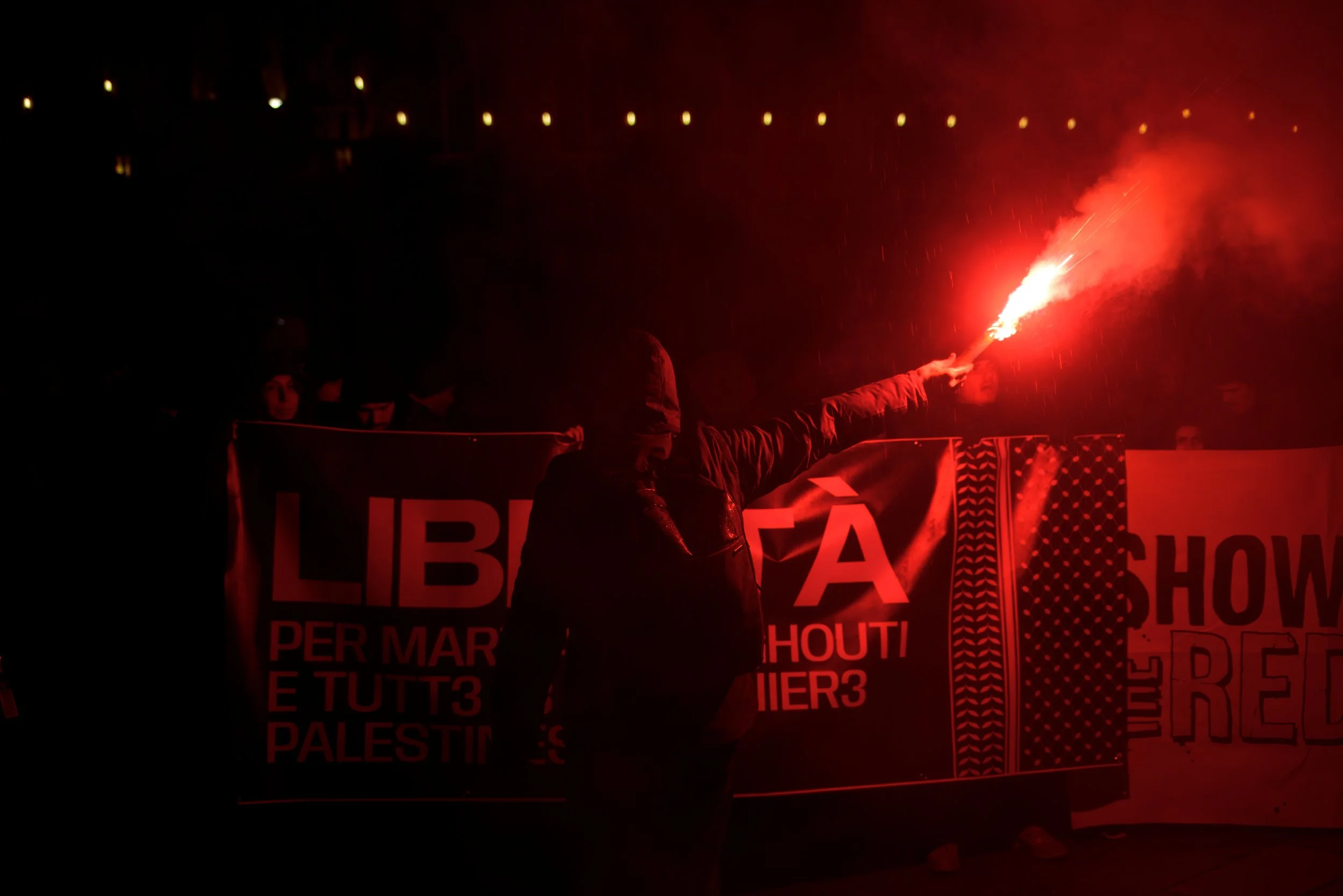Bologna, Italy – November 21, 2025: A hooded demonstrator raises a red flare in front of a banner calling for Palestinian freedom.