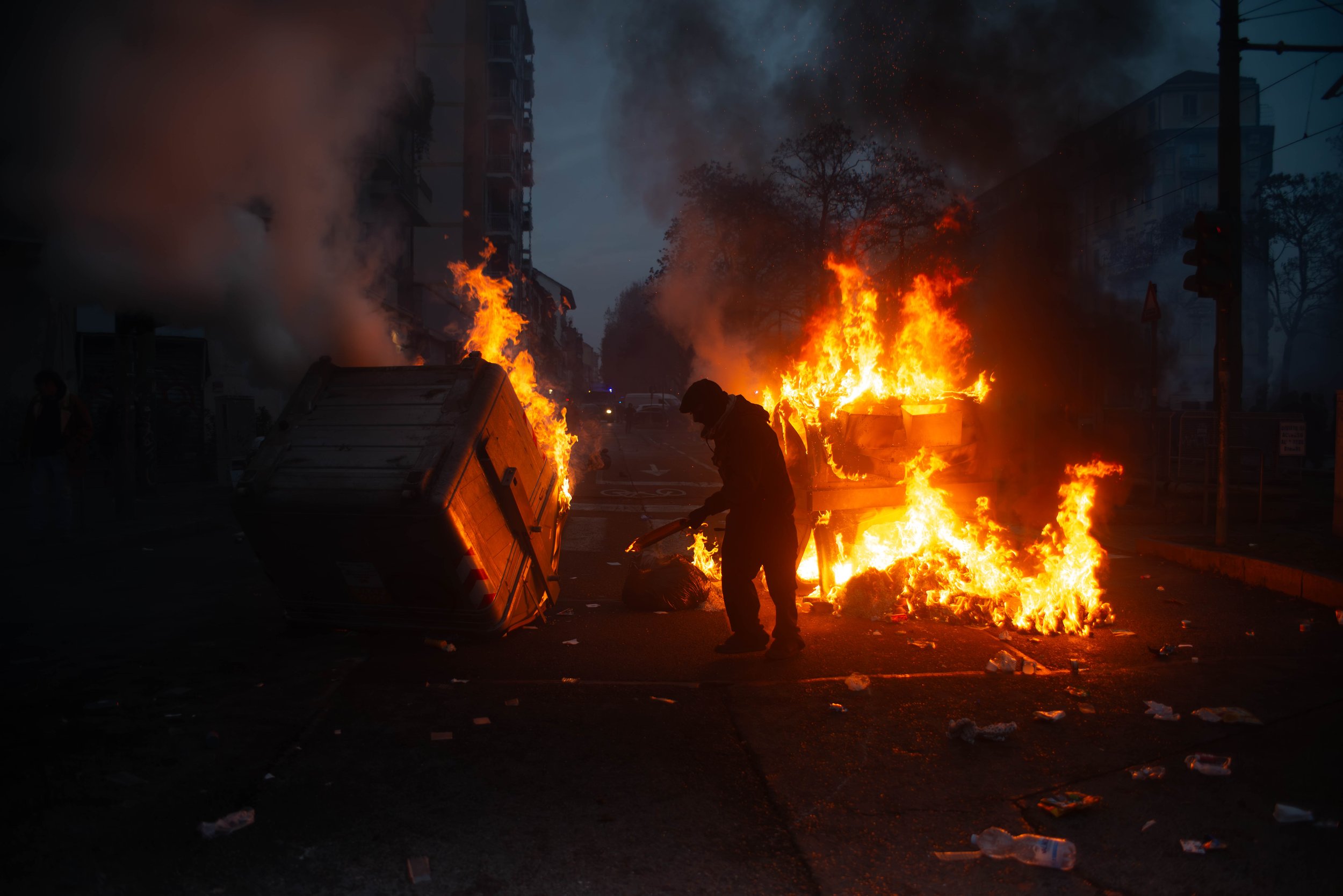 Turin, Italy – December 20, 2025: Flames from burning trash containers light up the street at dusk as a protester stands nearby during unrest following the eviction of the Askatasuna social center.