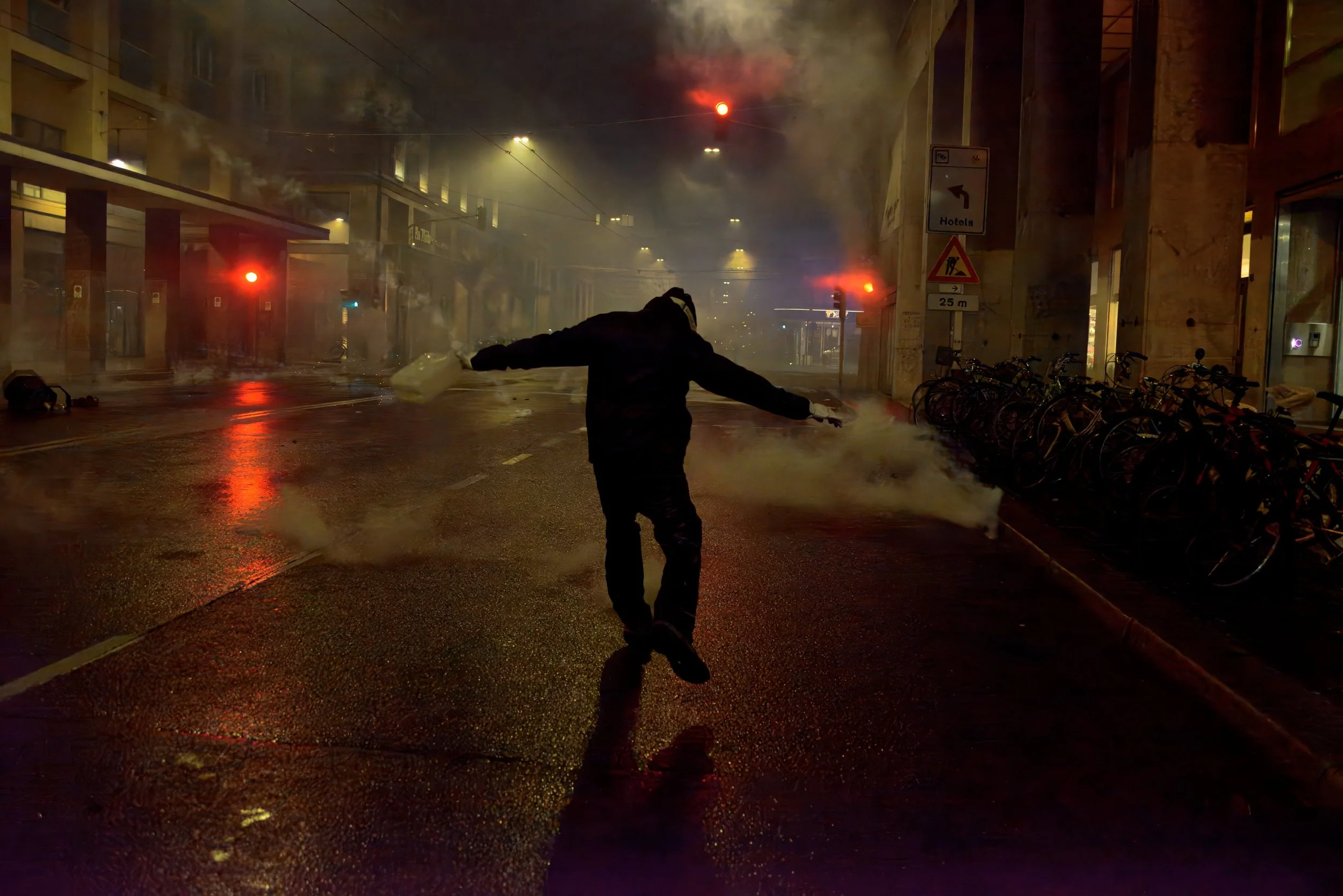 Bologna, Italy – November 21, 2025: A protester sprints down the middle of the road, arms wide open and body silhouetted against streetlights and traffic signals, cutting through tear gas and spray.
