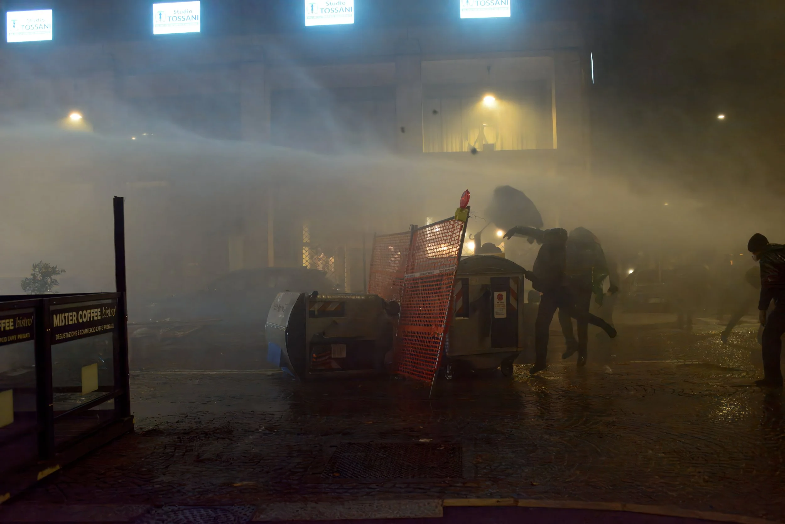 Bologna, Italy – November 21, 2025: Protesters repurpose dumpsters and metal barriers into makeshift barricades as water cannons sweep the street, turning the downtown confrontation over the game into a scene of urban trench warfare.