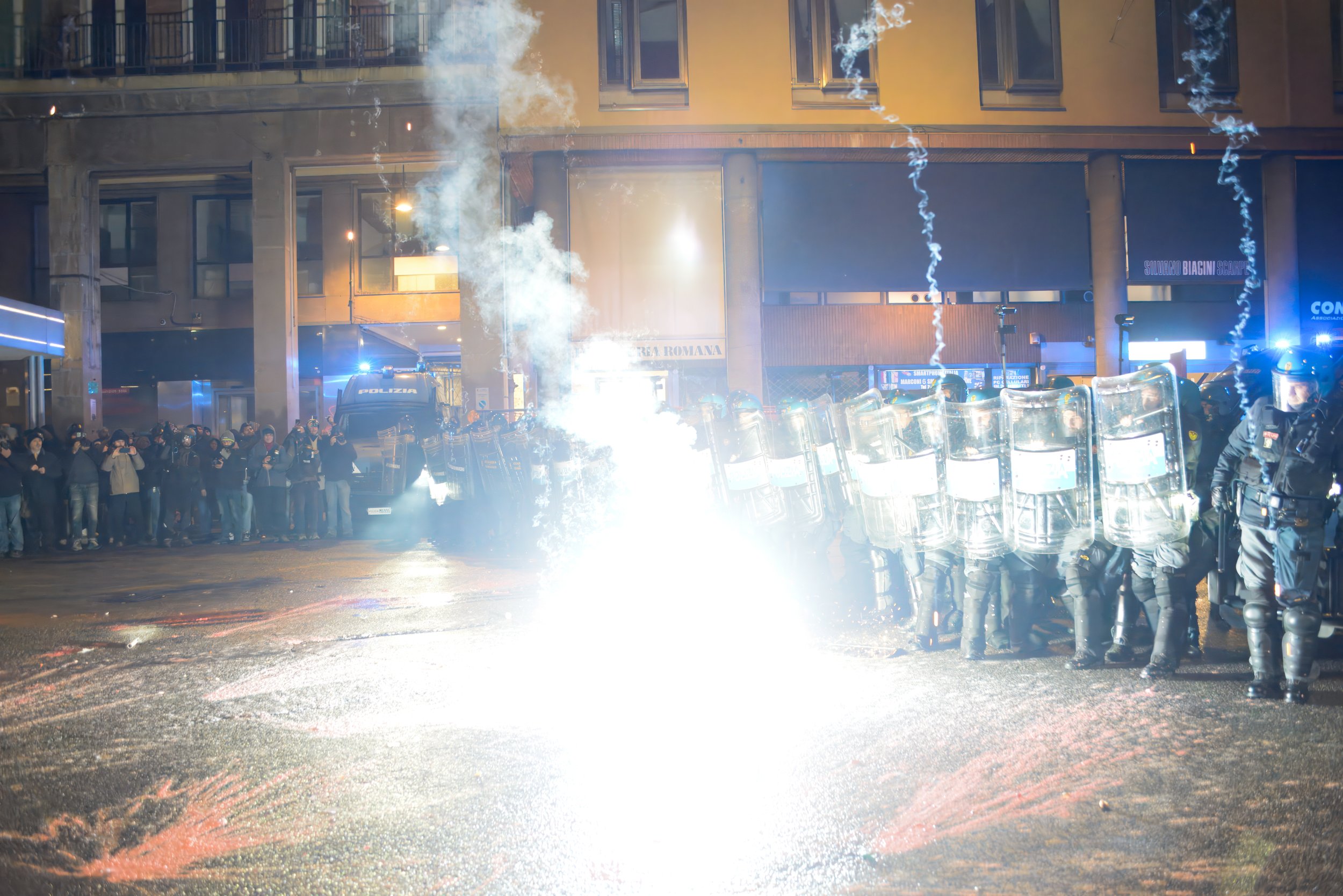 Bologna, Italy – November 21, 2025: A purple-white flash erupts on the asphalt just meters from the police cordon, illuminating shields, helmets and the faces of protesters across the street as the standoff escalates.