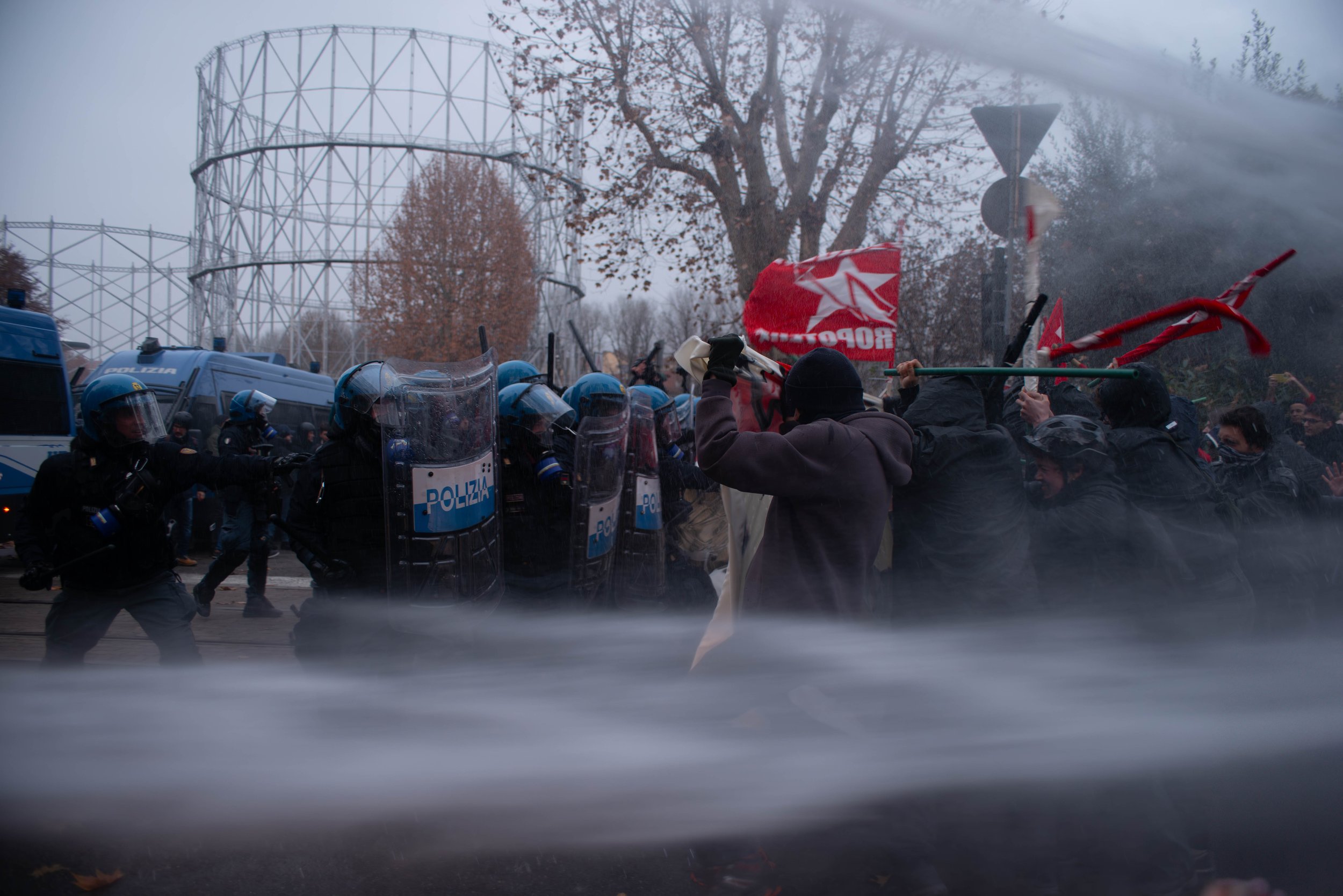 Turin, Italy – December 20, 2025: Riot police in helmets and shields clash with protesters brandishing red flags as powerful water cannons sweep across the frontline during demonstrations against the eviction of the Askatasuna social center.
