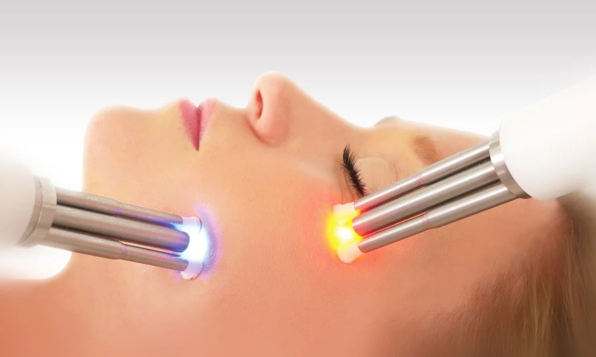Person receiving a facial laser treatment with two handheld laser devices emitting blue and red light.