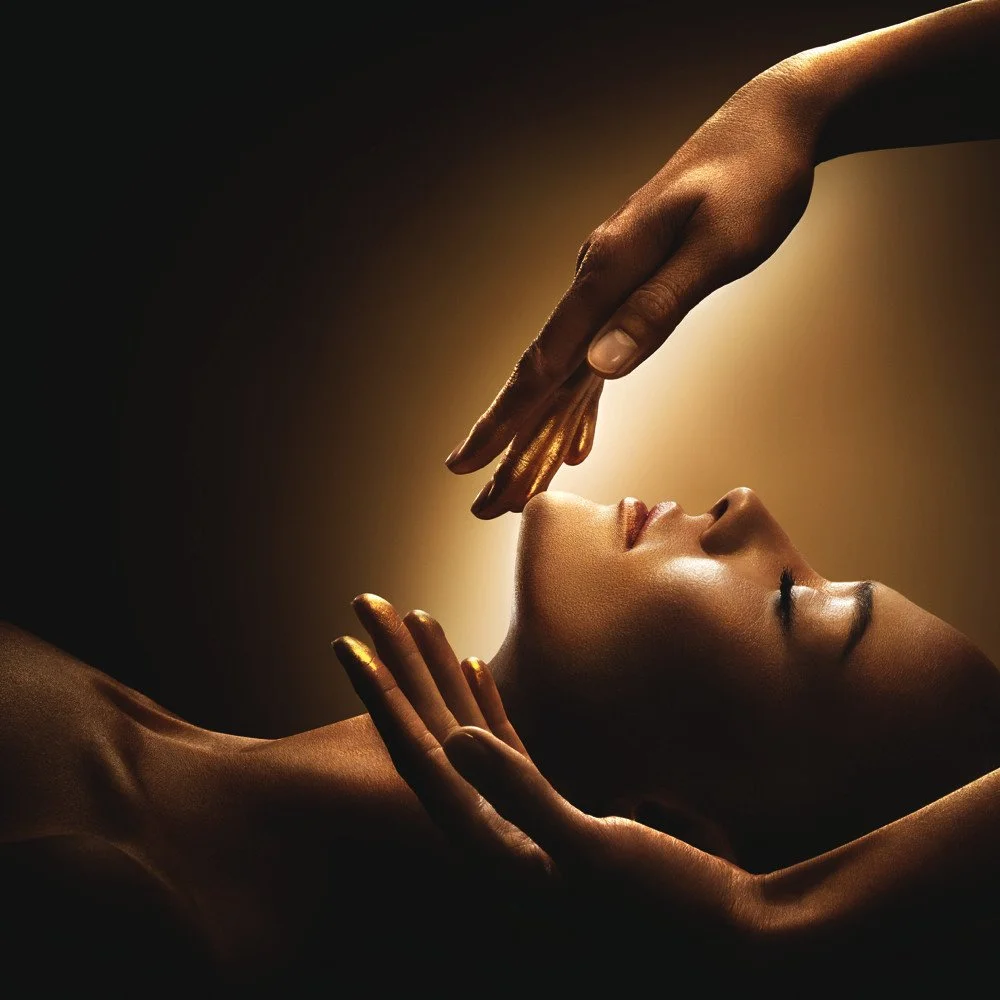 A person with closed eyes is lying down, with two hands gently touching their face and forehead, illuminated with warm lighting.