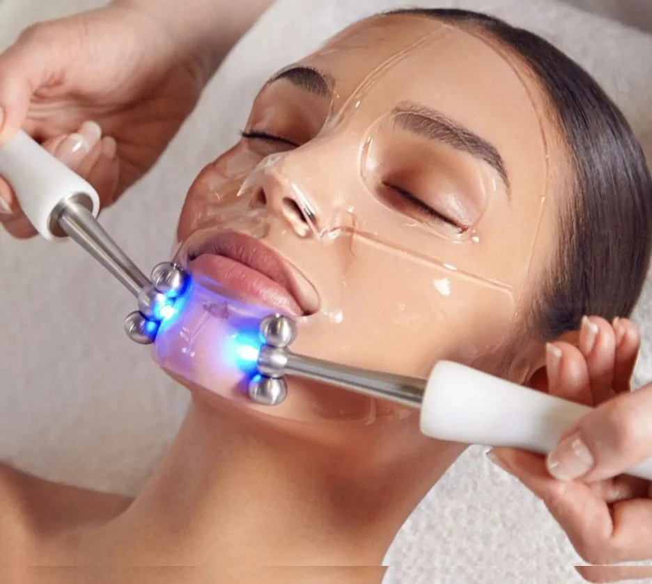 A woman receiving a facial treatment with a clear mask and blue LED light therapy, lying down with eyes closed.