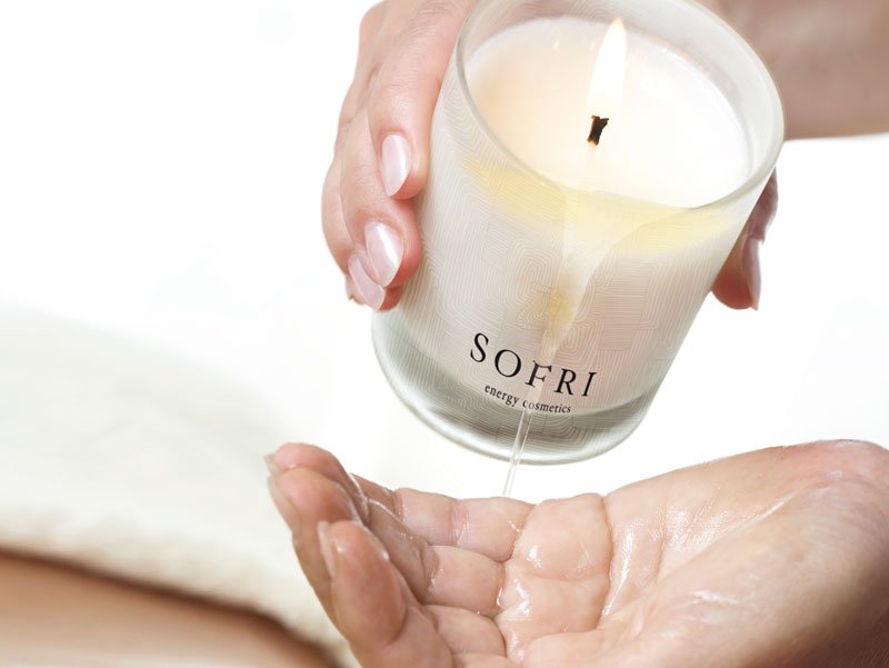 A white candle in a glass holder labeled 'SOFRI energy cosmetics' is being poured over a person's open palm.