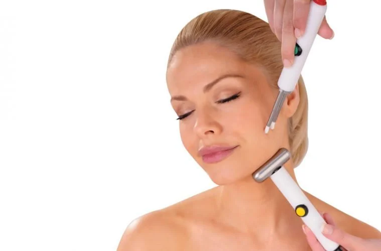 Woman with closed eyes receiving a facial treatment with a derma roller and a small device held by a practitioner.