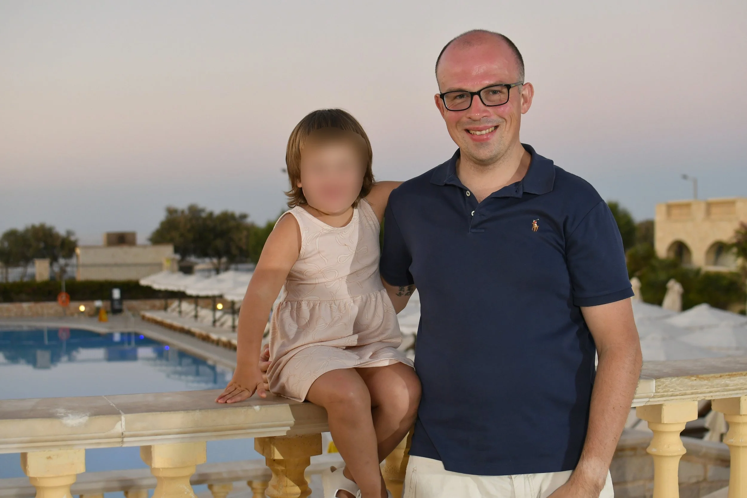 A man and a young girl are posing together outdoors near a swimming pool during sunset. The girl is sitting on a ledge with her arm around the man, both smiling at the camera.