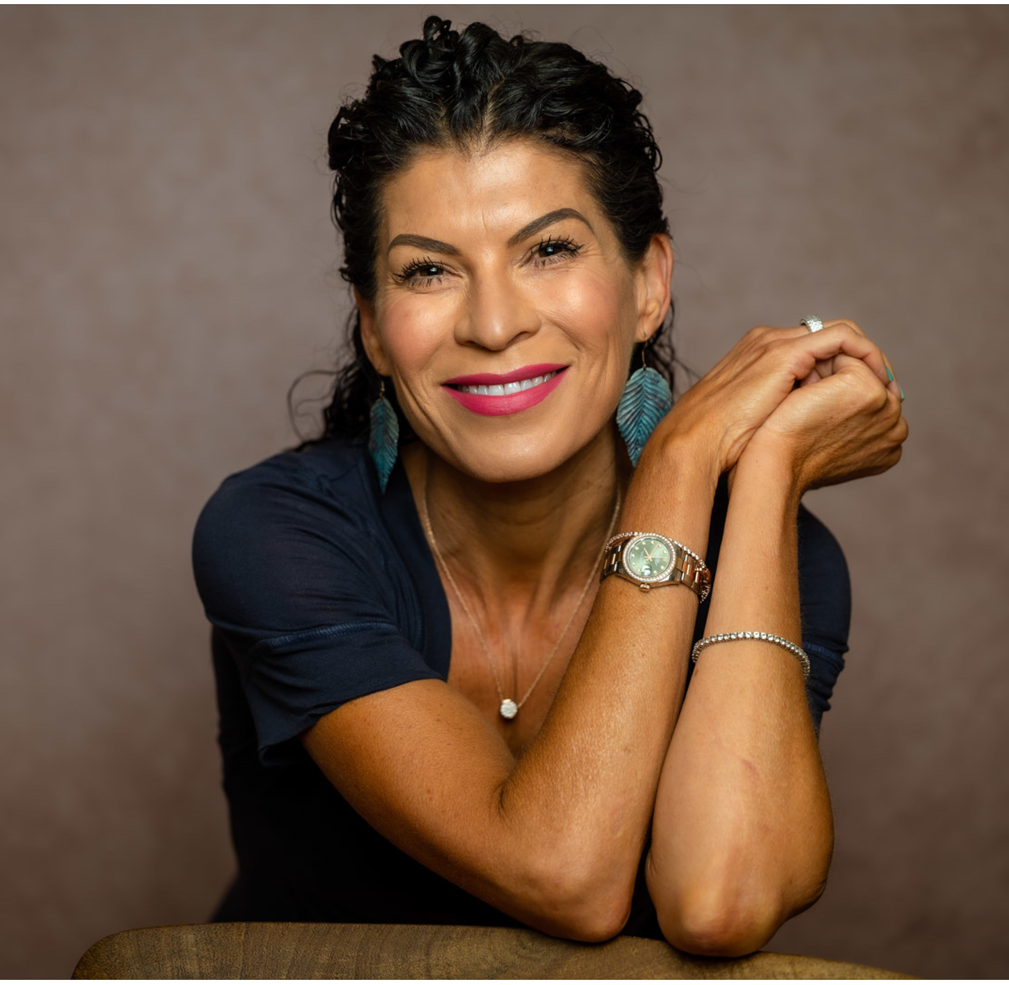 A smiling woman with dark curly hair, wearing turquoise earrings, a black top, and jewelry including a watch and bracelets, sitting with her arms crossed on a brown surface against a neutral background.