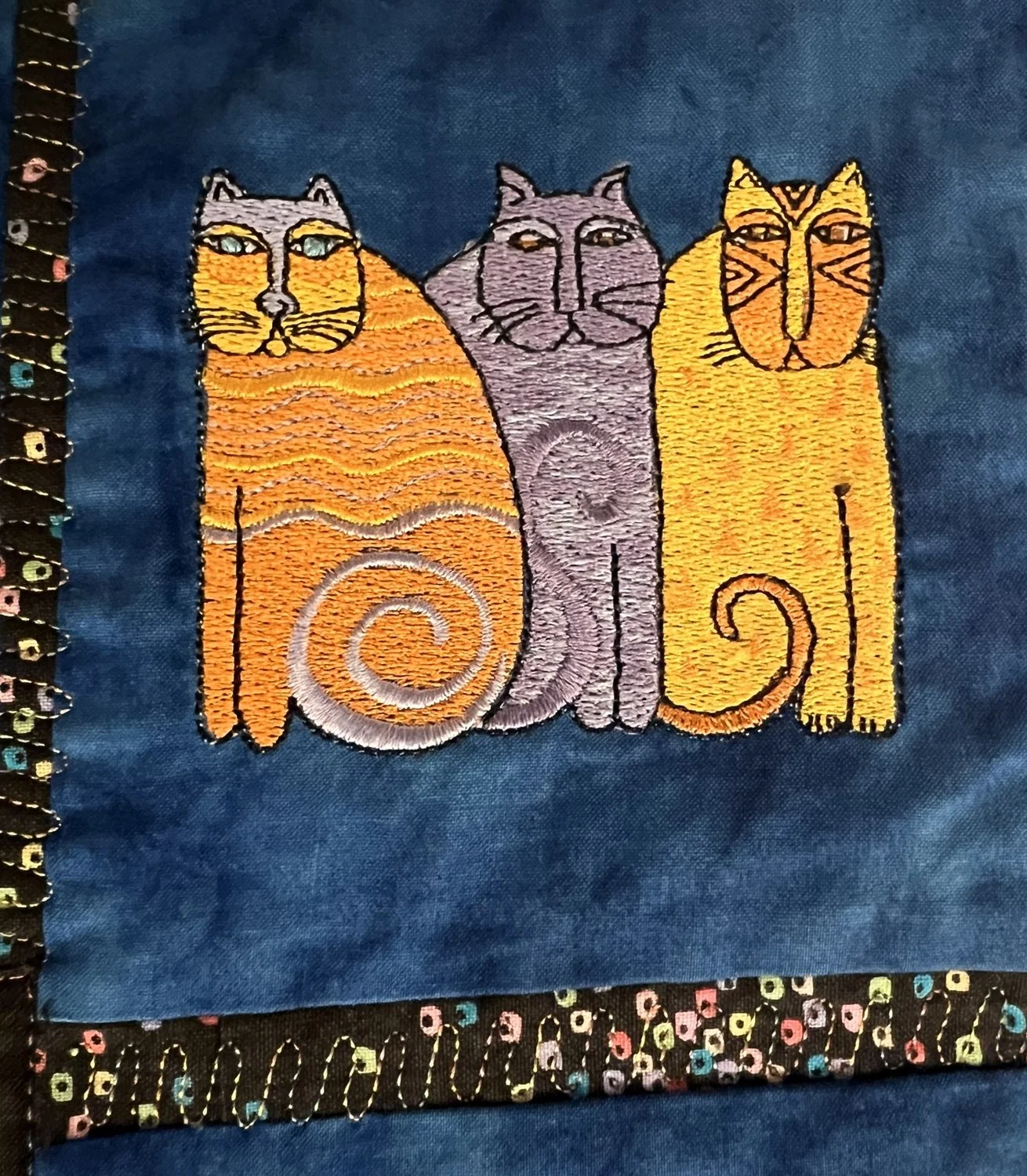 Embroidery of three stylized cats sitting side by side on dark fabric, with the left and right cats in orange and yellow, and the middle cat in purple.