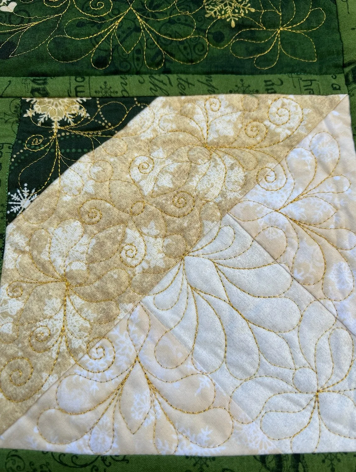 Close-up of a quilt with intricate gold stitching on a cream and beige fabric pattern, featuring swirling lines and leaf motifs.