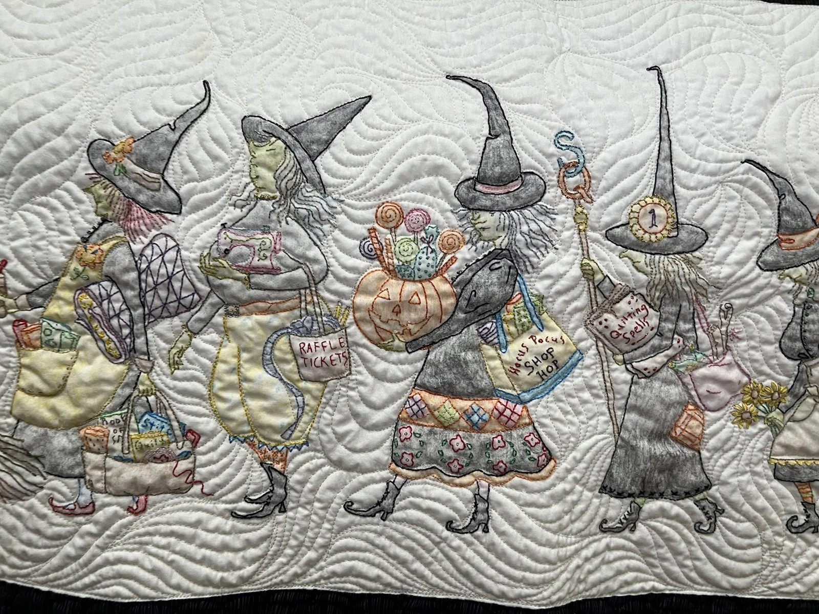 A quilt with embroidered witches carrying shopping bags and a pumpkin, all dressed in festive costumes.