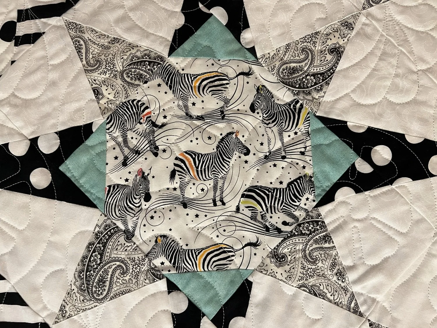 Close-up of a quilt with a central fabric square featuring multiple zebras, surrounded by various patterned fabrics in black, white, and teal colors.