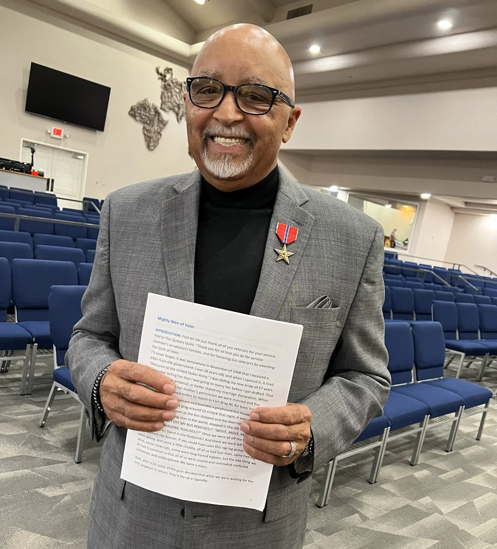 A man smiling, wearing glasses, a gray suit jacket with a black turtleneck, and a medal on his lapel, standing in an auditorium holding a sheet of paper.