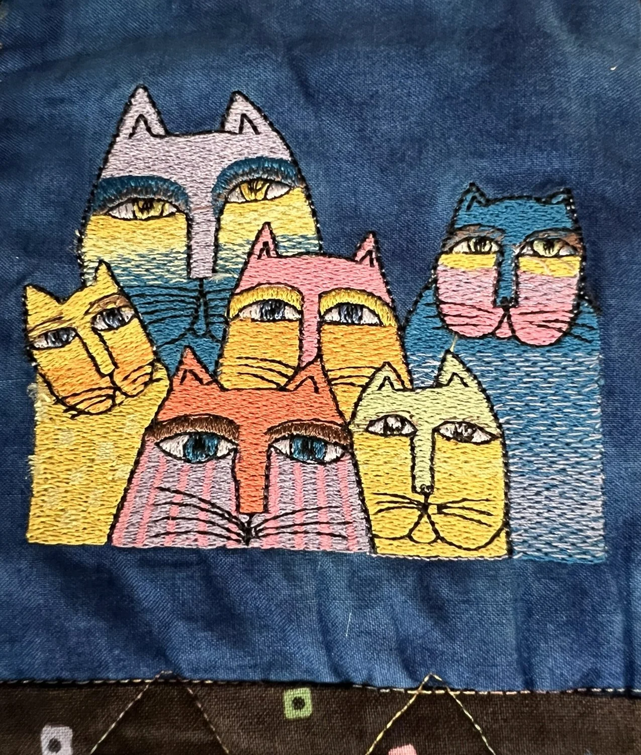 Colorful embroidery of seven cats with large eyes on blue fabric.