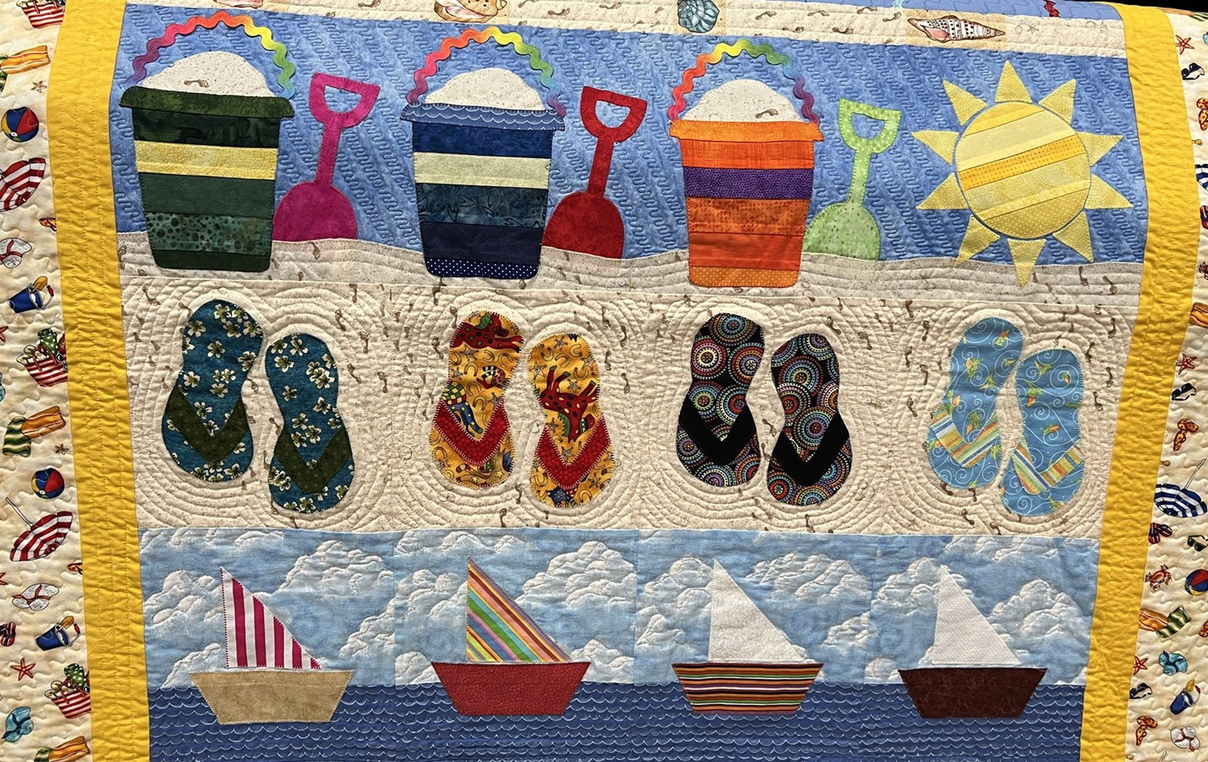 Colorful quilt depicting a beach scene with sand, flip-flops, pails, a sun, and boats on the water.