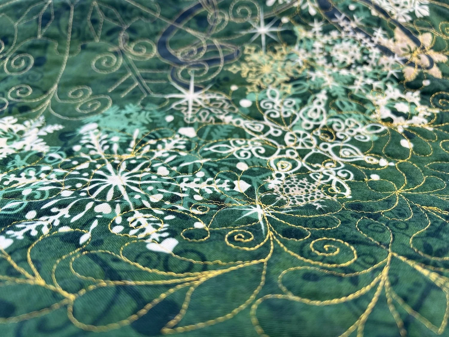 Close-up of a green fabric with intricate white and gold embroidered snowflake and swirl patterns.