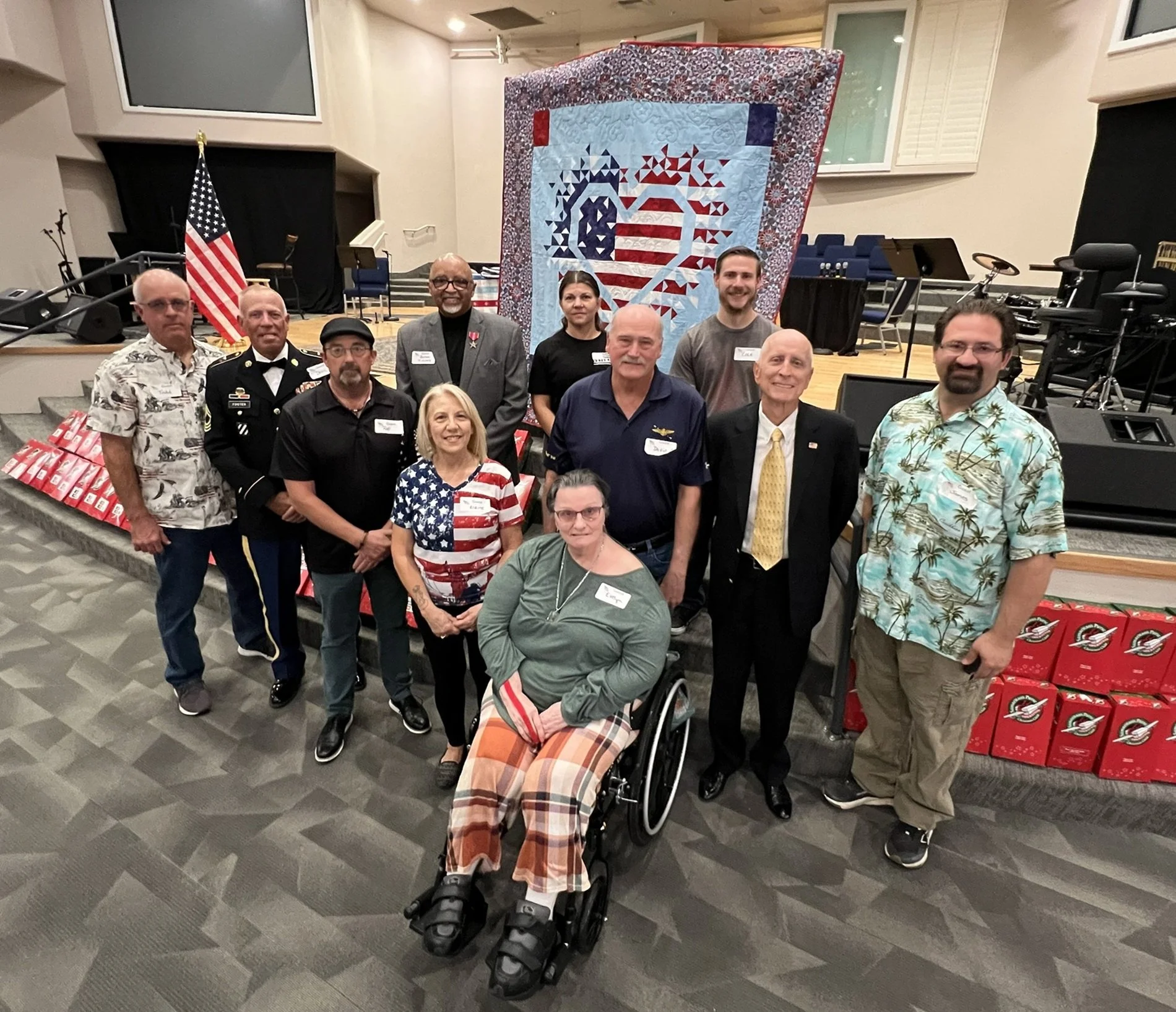 Group of people standing together at a patriotic event, with a quilt featuring an American flag and American eagle, American flags, and boxes of popcorn in the background.