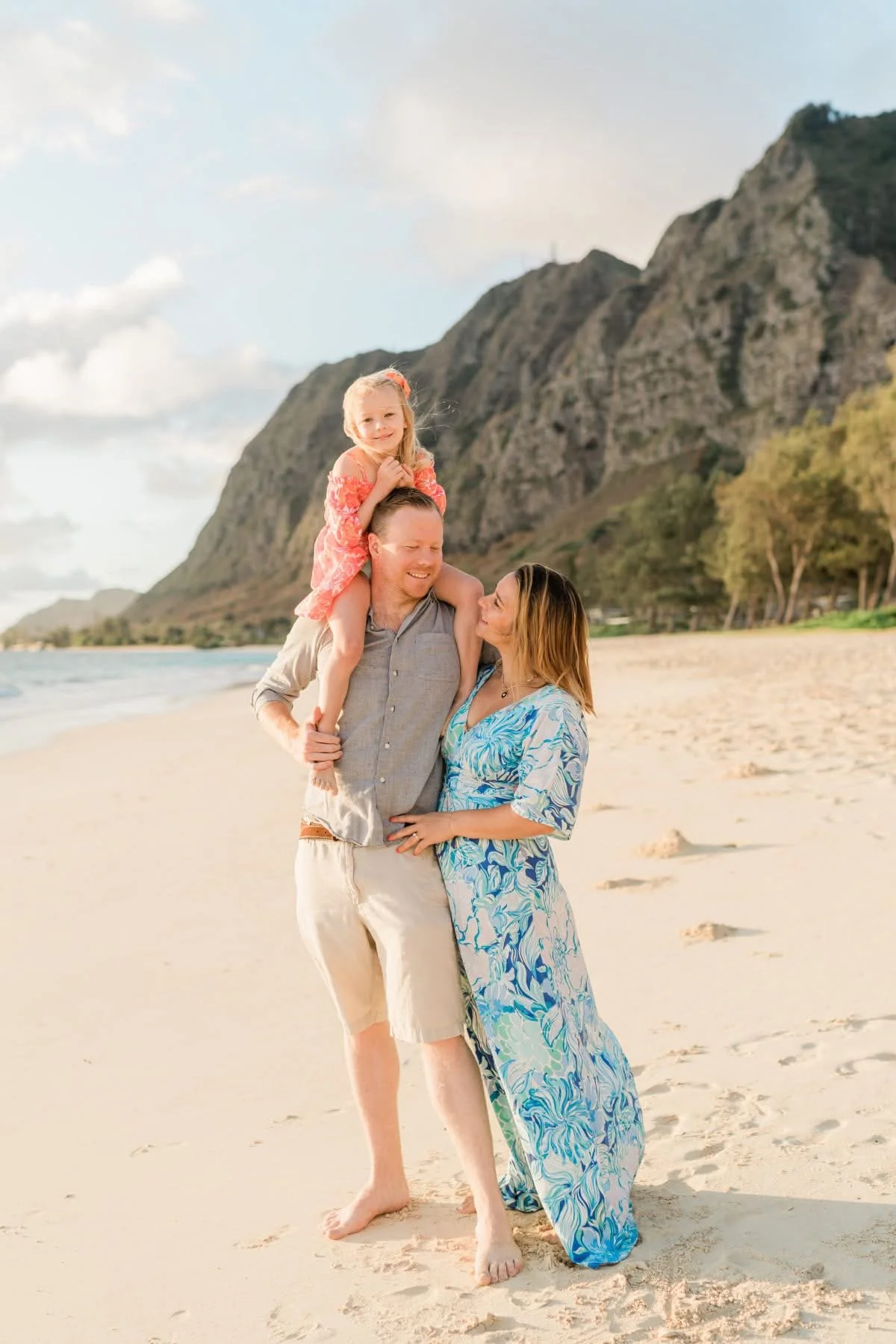 A family of three on a beach with mountains in the background. The man is holding a young girl on his shoulders, and the woman is standing beside them. They are smiling and enjoying a sunny day.